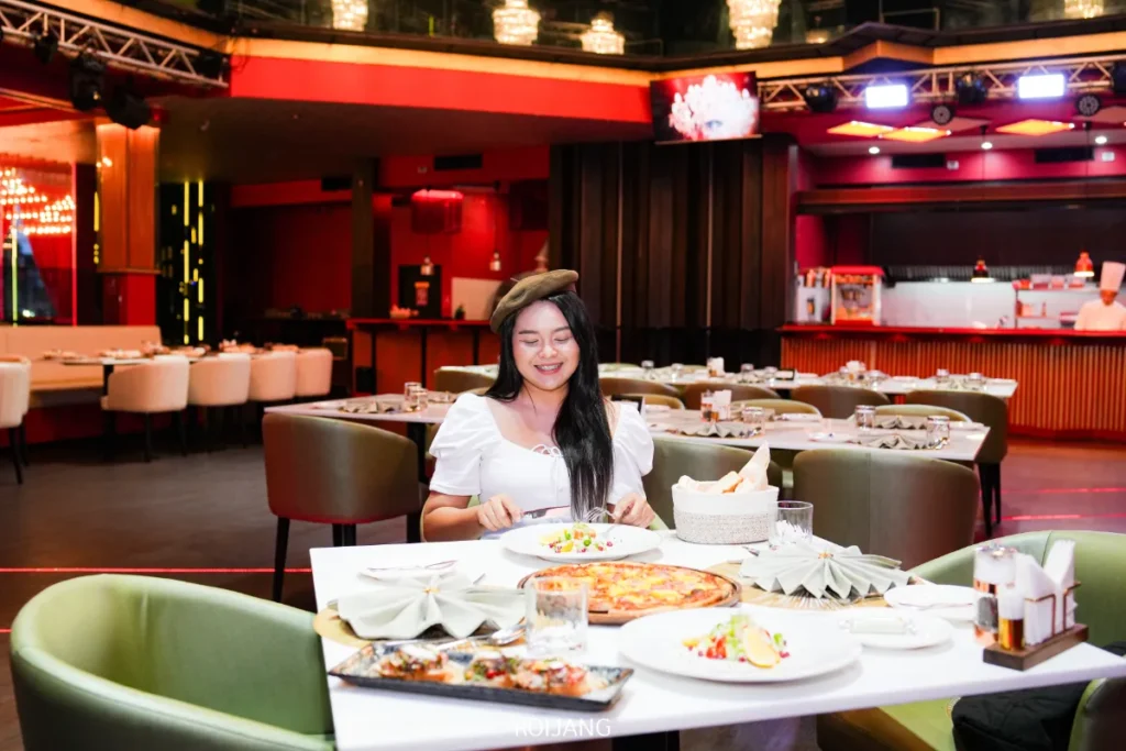ผู้หญิงคนหนึ่งนั่งอยู่คนเดียวที่โต๊ะในร้านอาหาร FLAME Phuket Town โดยมีพิซซ่า สลัด และเครื่องดื่มอยู่ตรงหน้าเธอ; โต๊ะว่างและแสงไฟอุ่น ๆ เต็มไปทั่วพื้นหลัง.