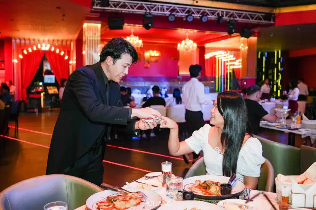 ชายในชุดสูทสีดำแสดงกลไพ่ให้กับหญิงสาวนั่งอยู่ที่ FLAME Phuket Town โดยมีผู้รับประทานอาหารคนอื่น ๆ มองเห็นได้อยู่เบื้องหลัง.
