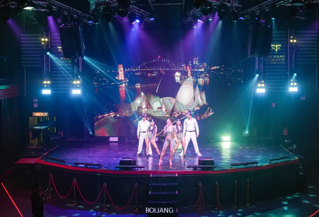นักร้องแสดงบนเวทีพร้อมกับนักเต้นสามคนในชุดกะลาสี ท่ามกลางแสงไฟที่สดใส และหน้าจอขนาดใหญ่ที่ฉายภาพเมืองยามค่ำคืน สื่อถึงชีวิตยามค่ำคืนที่เต็มไปด้วยพลังของ FLAME Phuket Town.