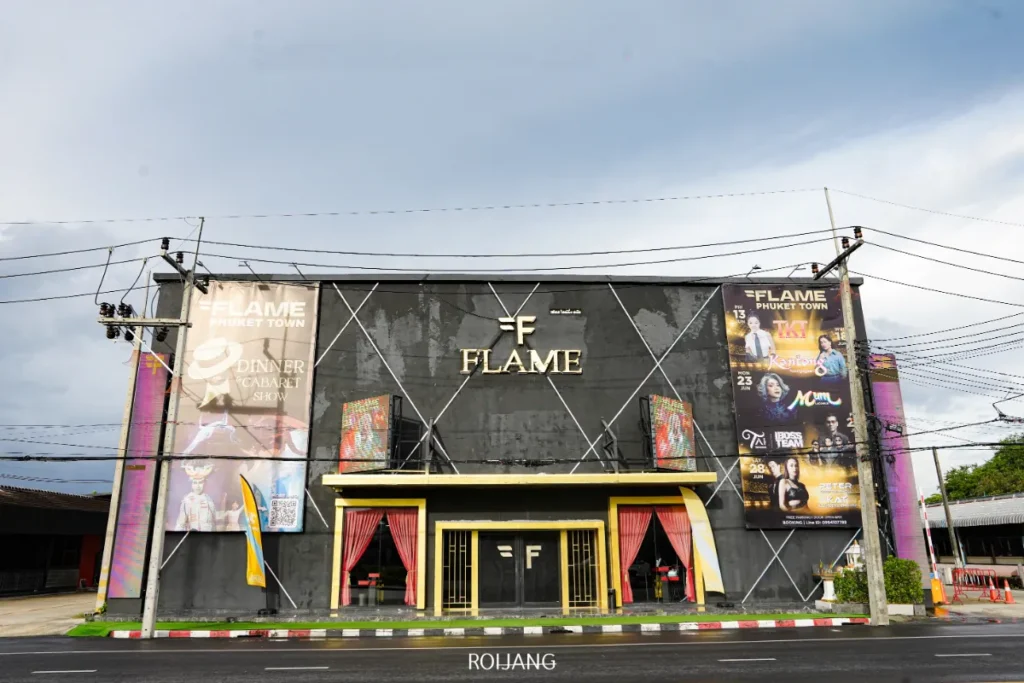 ด้านนอกของ FLAME Phuket Town nightclub มีป้ายโฆษณาโปรโมชั่นและทางเข้าที่ตกแต่งด้วยสีเหลือง ตั้งอยู่บนถนนที่มีสายไฟพาดผ่านเหนือศีรษะ.