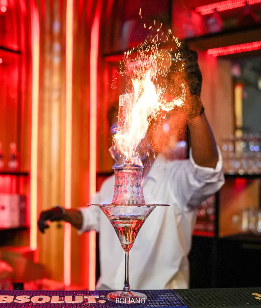 บาร์เทนเดอร์ที่ FLAME Phuket Town จุดไฟค็อกเทล ส่งเปลวไฟพุ่งขึ้นเหนือแก้วที่วางซ้อนกันในบาร์ที่มีแสงสลัวและแสงสีแดง.