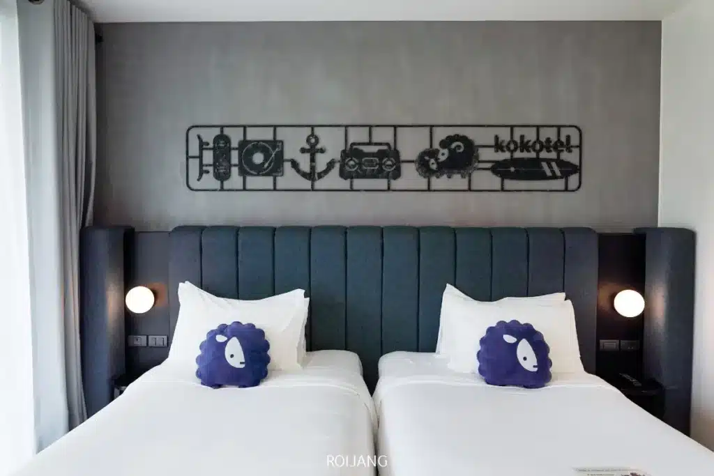 ห้องโรงแรม Kokotel ภูเก็ตในยาง ที่มีเตียงคู่สองเตียงพร้อมผ้าปูที่นอนสีขาวและหมอนรูปสัตว์สีน้ำเงิน มีศิลปะบนผนังที่ได้แรงบันดาลใจจากแอบแอ และโคมไฟข้างเตียงที่มีสไตล์ตั้งอยู่ทั้งสองข้าง.