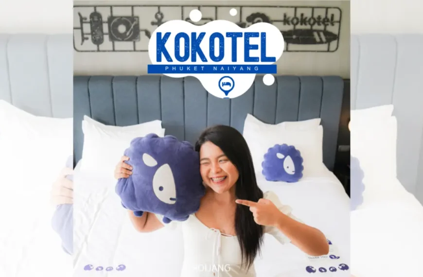 ผู้หญิงคนหนึ่งนั่งอยู่บนเตียงในห้องโรงแรม ยิ้มและถือหมอนสีน้ำเงินที่มีหน้าแกะการ์ตูน โลโก้ของ Kokotel Phuket Naiyang อยู่บนหัวเตียง สร้างบรรยากาศที่อบอุ่นและสบายๆ.
