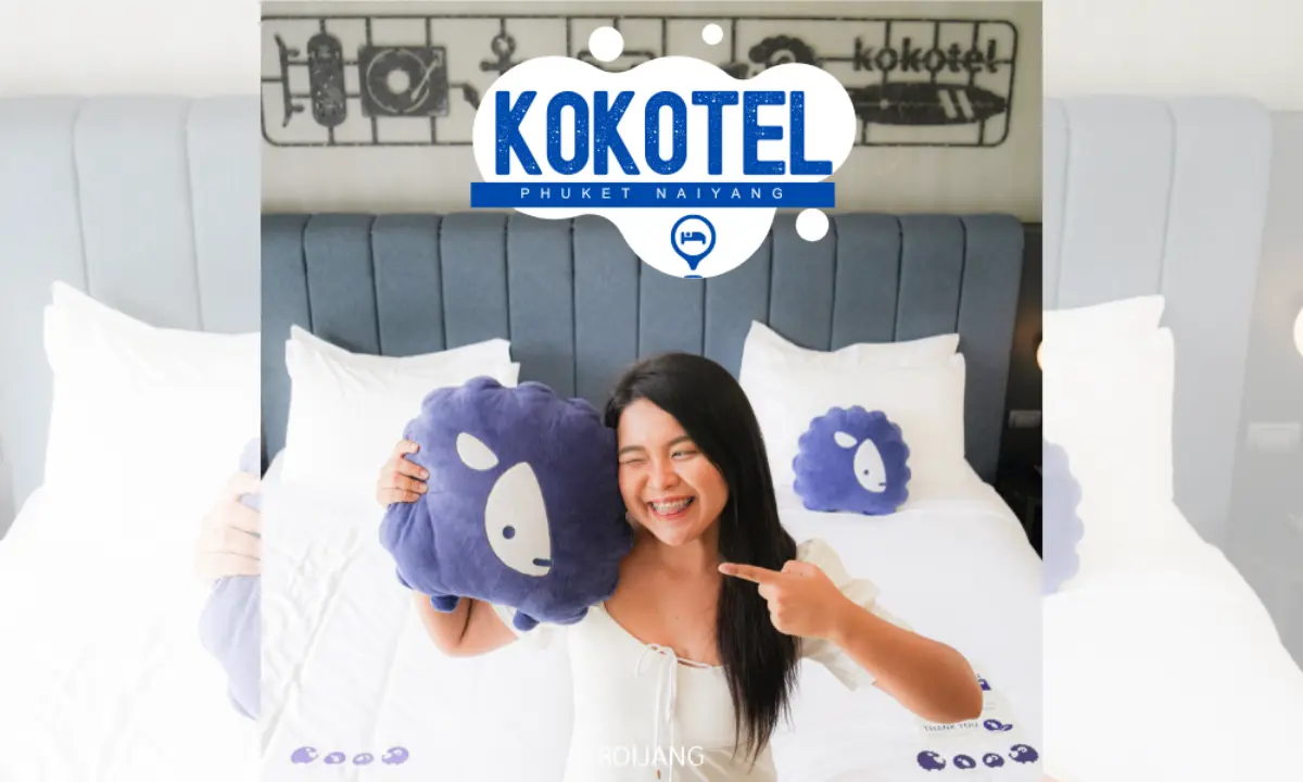 ผู้หญิงคนหนึ่งนั่งอยู่บนเตียงในห้องโรงแรม ยิ้มและถือหมอนสีน้ำเงินที่มีหน้าแกะการ์ตูน โลโก้ของ Kokotel Phuket Naiyang อยู่บนหัวเตียง สร้างบรรยากาศที่อบอุ่นและสบายๆ.