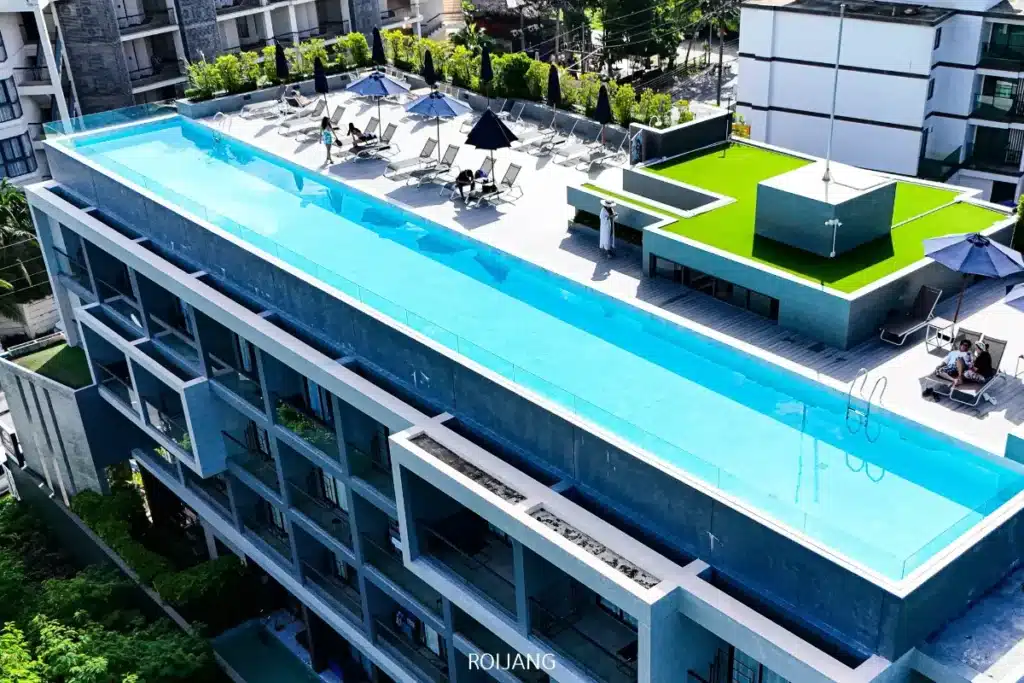 สระว่ายน้ำบนดาดฟ้าที่มีผนังใสทอดยาวตลอดความยาวของอาคาร  Kokotel ภูเก็ตในยาง สมัยใหม่ ที่ซึ่งผู้อยู่อาศัยสามารถผ่อนคลายบนเก้าอี้อาบแดดใต้ร่มหรือเพลิดเพลินกับพื้นที่สนามหญ้าสีเขียวเพื่อบรรยากาศสบายๆ แบบแอบแอ.