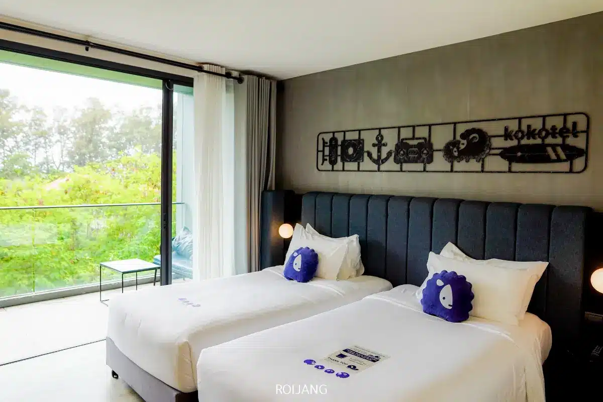 ห้องพักโรงแรมสมัยใหม่พร้อมเตียงคู่สองเตียง ผ้าปูที่นอนสีขาว หมอนนุ่มสีม่วง และงานศิลปะบนผนังเหนือหัวเตียง มอบการพักผ่อนที่เงียบสงบ ในขณะที่ระเบียงให้ทัศนียภาพที่สดชื่นของพื้นที่สีเขียว—เหมาะสำหรับการผ่อนคลายหรือเพลิดเพลินกับช่วงเวลาของ แอบแอ.