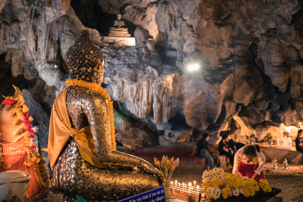 พระพุทธรูปทองคำที่ห่มด้วยผ้าสี ที่เที่ยวเชียงดาว เหลืองนั่งอยู่ภายในถ้ำ ล้อมรอบด้วยเครื่องบูชา เทียน และผู้มาเยือนที่กำลังสวดมนต์—เป็นประสบการณ์อันสงบเงียบของสถานที่ท่องเที่ยวเชียงดาว.