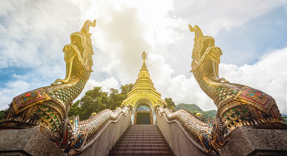 บันไดวัดที่ประดับประดาอย่างวิจิตร มีรูปปั้นงูยักษ์นาคาสีทองขนาดใหญ่ ที่เที่ยวเชียงดาว สองตัวขนาบข้าง นำทางขึ้นไปยังเจดีย์สีทองที่ตั้งตระหง่านท่ามกลางท้องฟ้าที่มีเมฆครึ้ม—เป็นสถานที่ที่ไม่ควรพลาดสำหรับผู้ที่มาเยือนแหล่งท่องเที่ยวเชียงดาว.