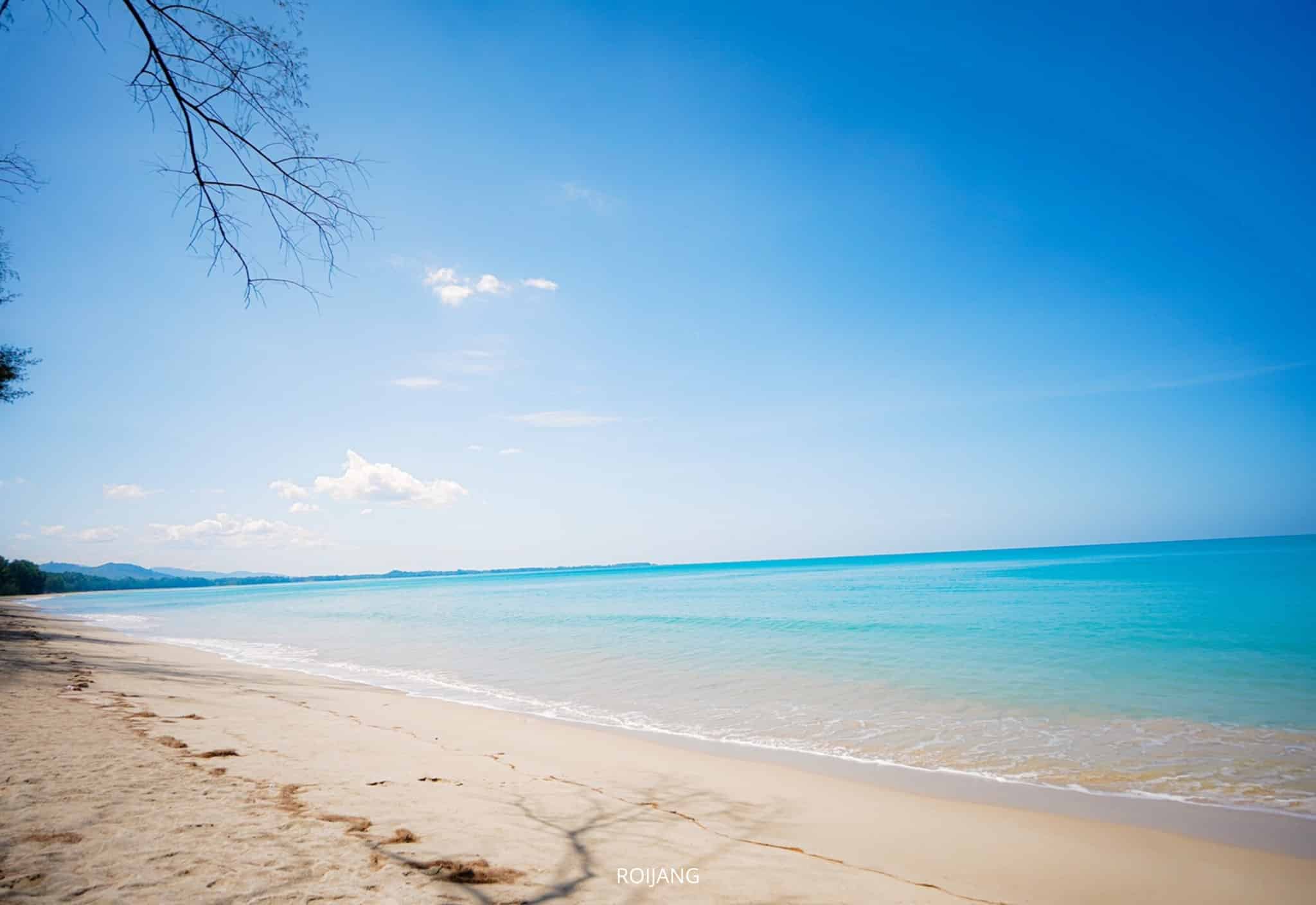 ภาพชายหาดที่เงียบสงบพร้อมน้ำทะเลสีฟ้าใสสะอาด ชายหาดที่ปกคลุมด้วยทรายสีอ่อน และท้องฟ้าสีครามที่มีเมฆกระจายอยู่เป็นจุด ๆ กิ่งไม้ที่บางตาโอบล้อมมุมซ้ายบนของภาพ ทำให้เห็นเสน่ห์ที่เงียบสงบของ ที่เที่ยวตะกั่วป่า