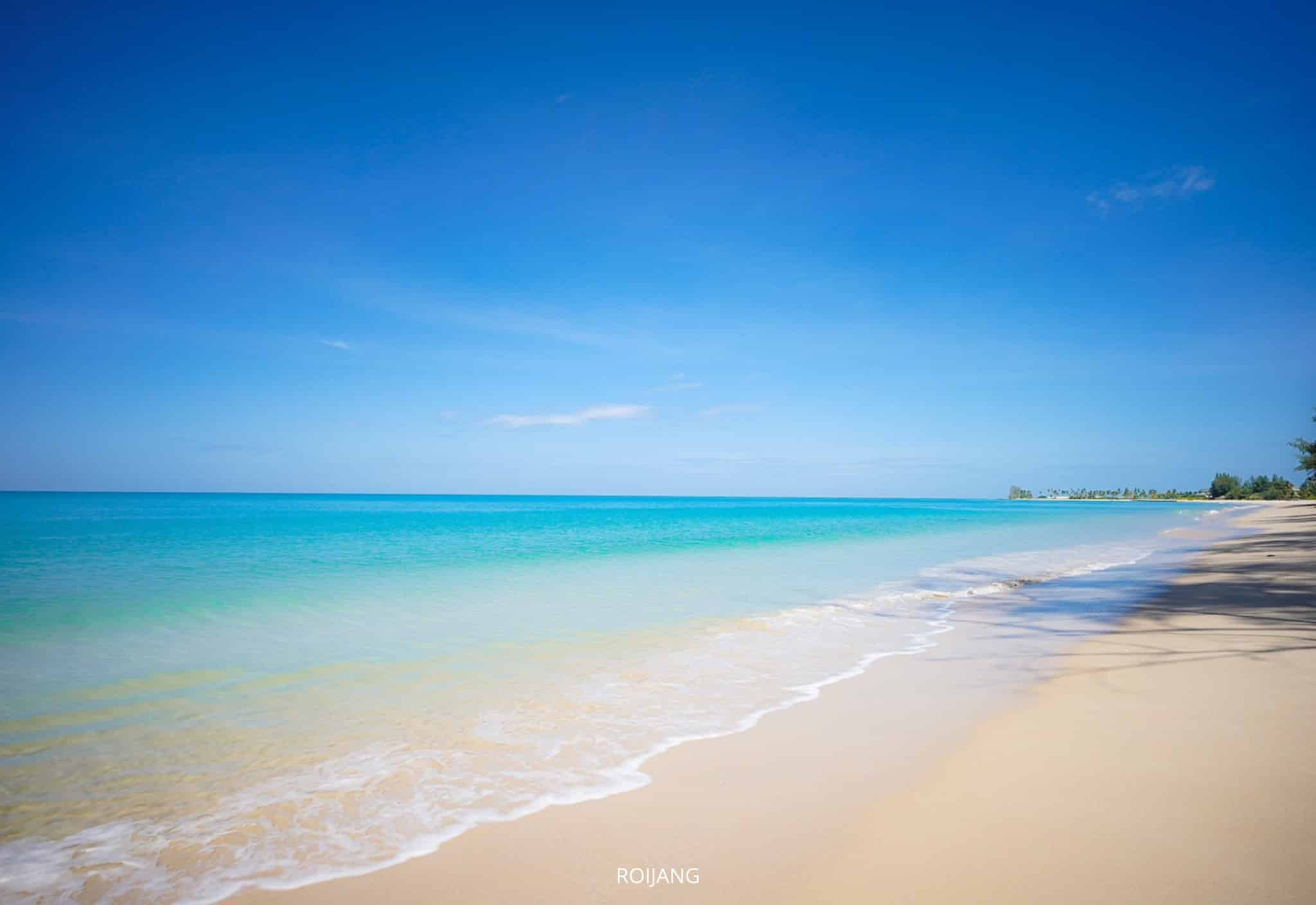 ท้องฟ้าสีฟ้าใสเหนือทะเลสีฟ้าครามที่สงบ คลื่นเบาๆ ซัดเข้าหาชายหาดทราย และต้นไม้ห่างๆ ตามแนวชายฝั่งด้านขวา—ภาพที่ชวนให้หลงใหลซึ่งเป็นลักษณะเฉพาะของ ที่เที่ยวตะกั่วป่า