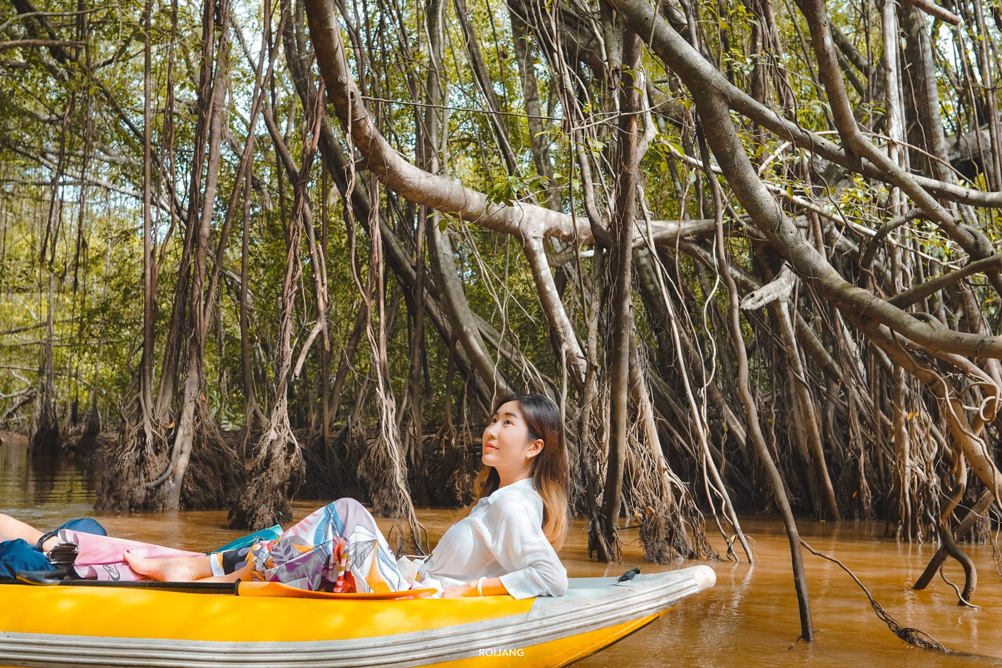 ผู้หญิงคนหนึ่งกำลังผ่อนคลายบนเรือคายัคสีเหลืองในแม่น้ำที่ล้อมรอบด้วยป่าชายเลนหนาแน่นที่มีรากโผล่ขึ้นมาเพลิดเพลินกับความงามตามธรรมชาติของ ที่เที่ยวตะกั่วป่า.