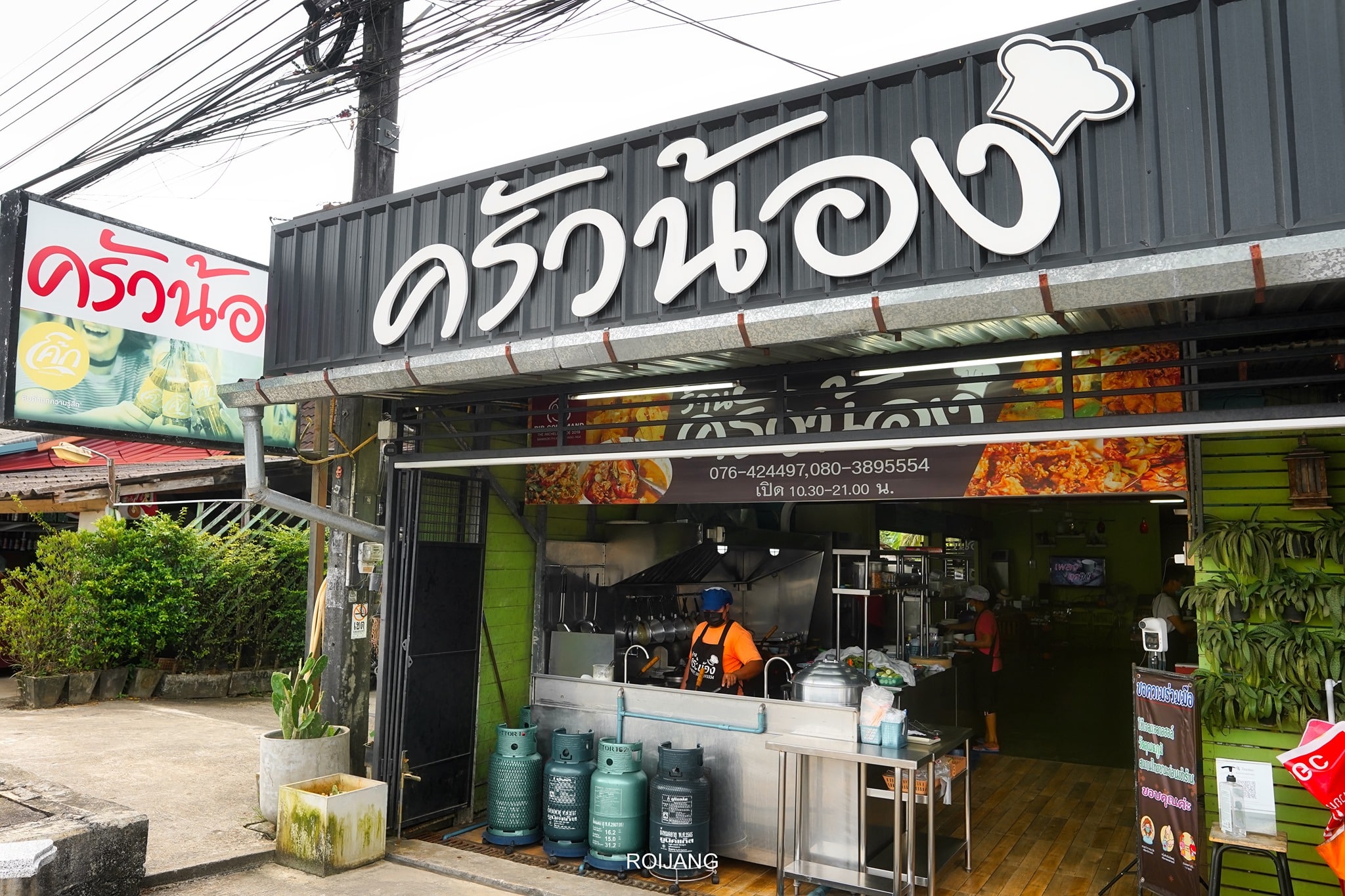 ภาพถนนหน้าร้านอาหารไทยขนาดเล็ก มีป้ายตัวอักษรไทยขนาดใหญ่ ถังแก๊สตั้งอยู่ด้านนอก และคนกำลังทำงานอยู่ใกล้ทางเข้า—หนึ่งในร้านเด็ดที่คุณอาจพบได้ขณะสำรวจ ที่เที่ยวตะกั่วป่า