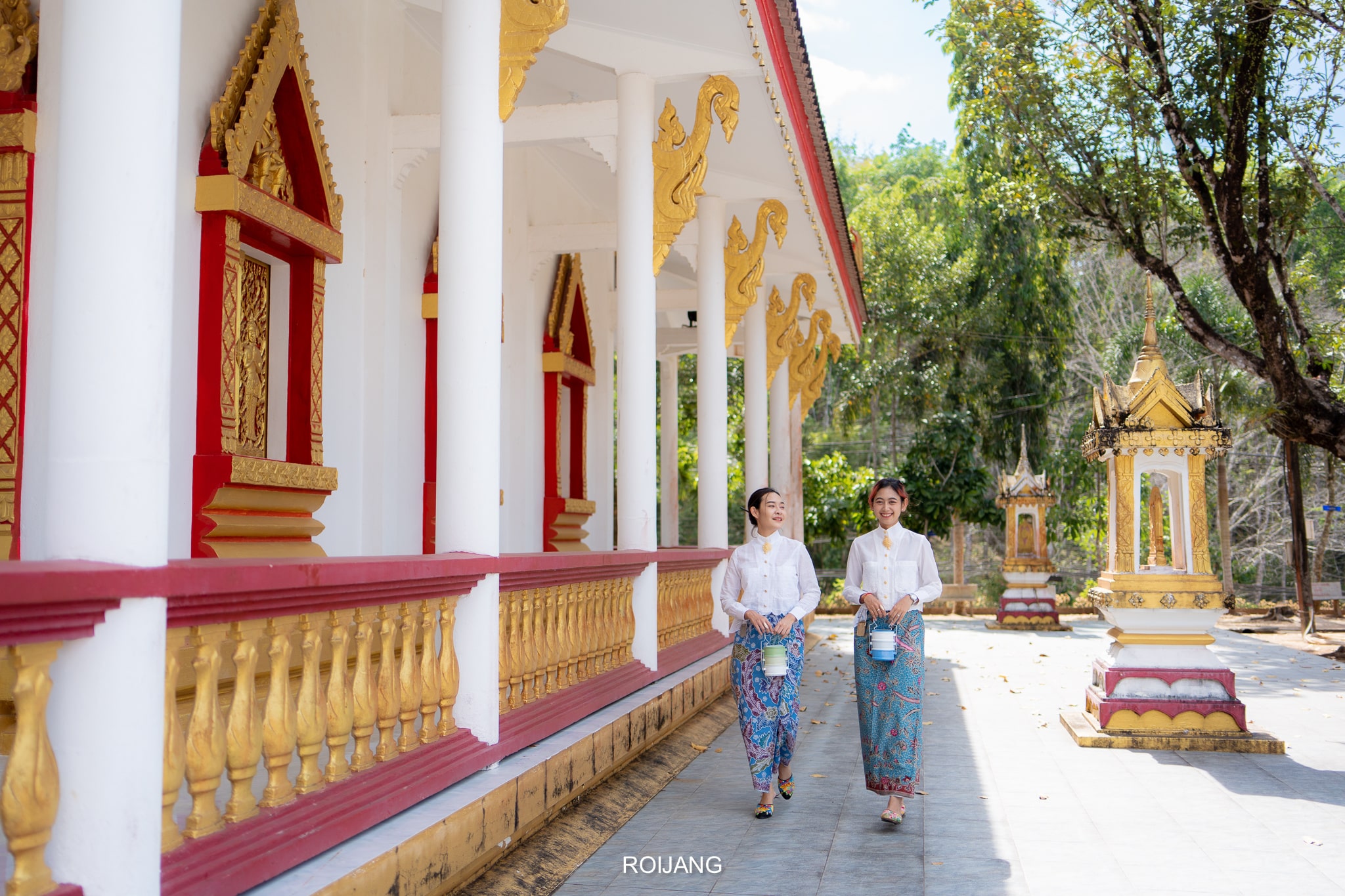 ผู้หญิงสองคนในชุดประจำชาติเดินไปตามด้านข้างของวัดที่มีเสาประดับประดาและตกแต่งด้วยทองคำในวันที่แดดจ้า สะท้อนถึงจิตวิญญาณที่สดใสของ ที่เที่ยวตะกั่วป่า.