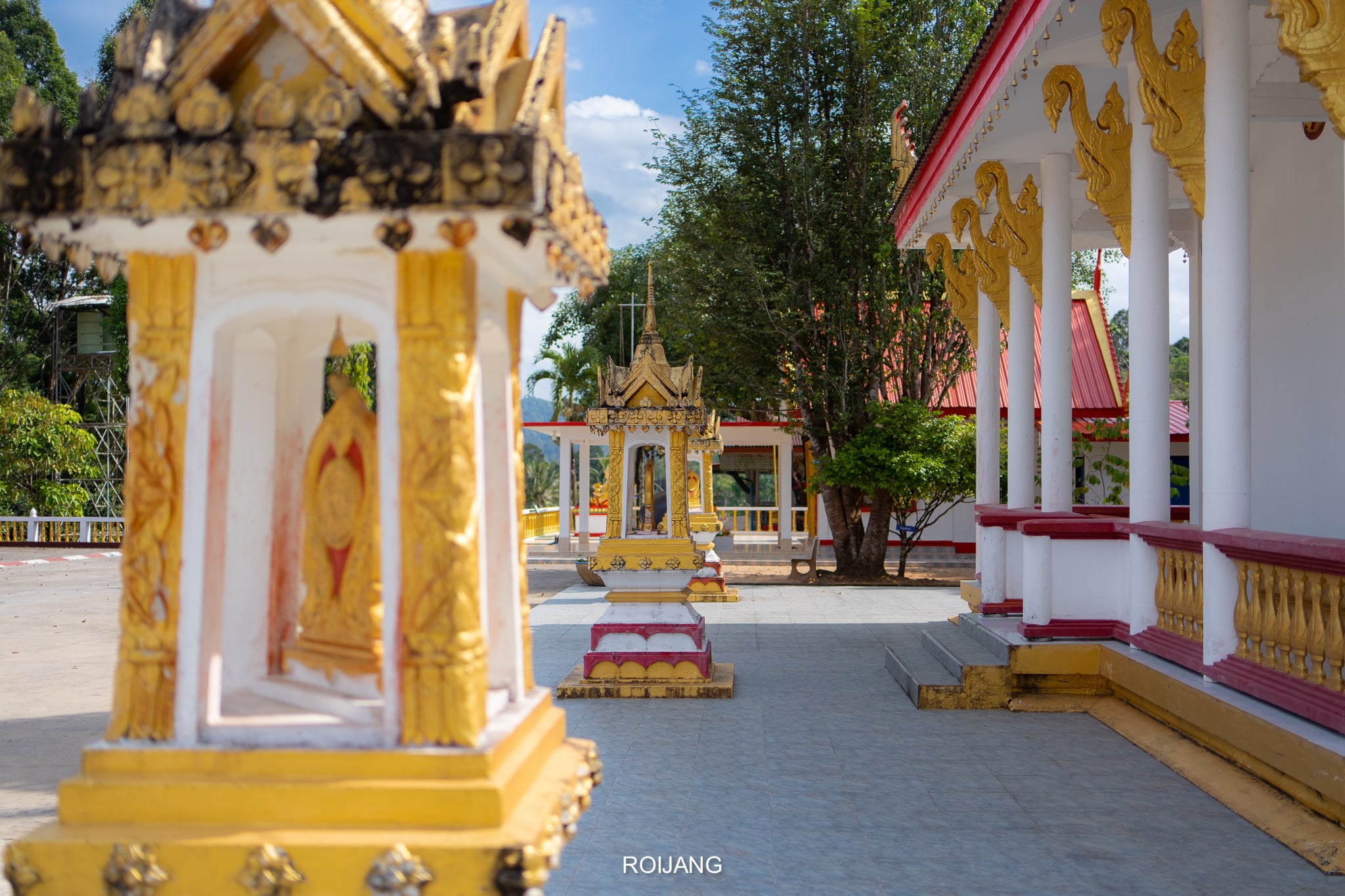 ลานวัดพุทธที่มีศาลทองคำและสีขาวประดับประดาอย่างสวยงาม เสาประดับตกแต่ง และต้นไม้ในฉากหลังภายใต้ท้องฟ้าแจ่มใส— สถานที่ท่องเที่ยวตะกั่วป่า ที่น่าเชิญชวนในตะกั่วป่าสำหรับผู้ที่แสวงหาความสงบและความงามทางวัฒนธรรม.