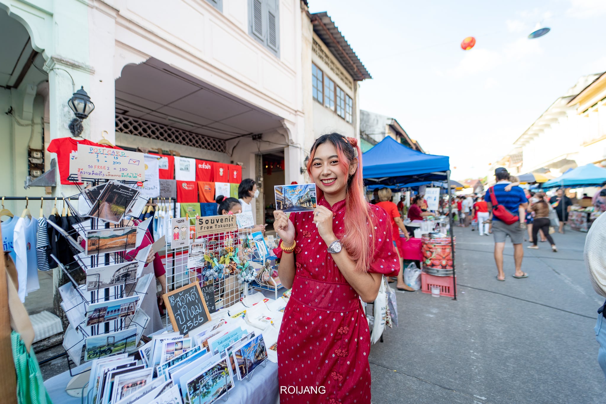 ผู้หญิงในชุดเดรสสีแดงยืนอยู่ที่แผงขายของในตลาดกลางแจ้ง ถือโปสการ์ดไว้ในมือ ล้อมรอบด้วยชั้นวางของที่ระลึกและโปสการ์ด—ภาพที่สดใสซึ่งสะท้อนเสน่ห์ของ ที่เที่ยวตะกั่วป่า.