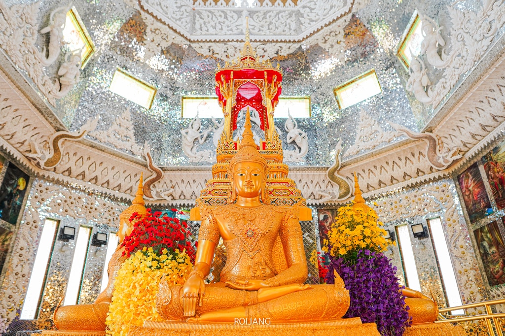 พระพุทธรูปทองคำประทับนั่งบนฐานที่ประดับตกแต่งอย่างวิจิตร ล้อมรอบด้วยพวงมาลัยดอกไม้สีสันสดใส ภายในวิหารที่ตกแต่งอย่างหรูหราด้วยกระจกเงา—เป็นไฮไลท์ที่ไม่ควรพลาดใน สถานที่ท่องเที่ยวตะกั่วป่า