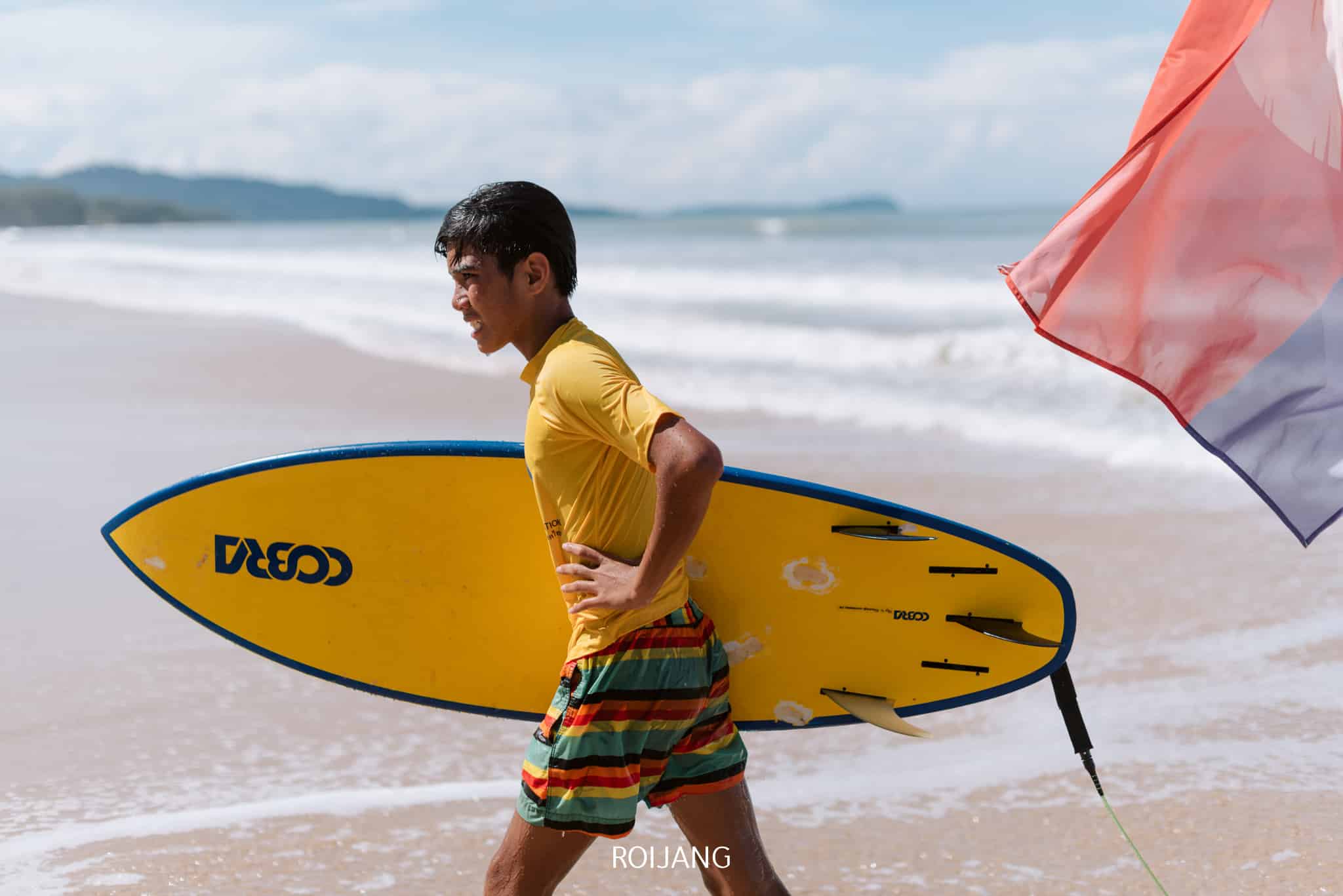 ชายคนหนึ่งสวมเสื้อเชิ้ตสีเหลืองและกางเกงขาสั้นลายทางถือกระดานโต้คลื่นสีเหลืองเดินไปตามชายหาดที่เต็มไปด้วยทราย ณ สถานที่ท่องเที่ยวตะกั่วป่า โดยมีคลื่นและธงปรากฏอยู่เบื้องหลัง.