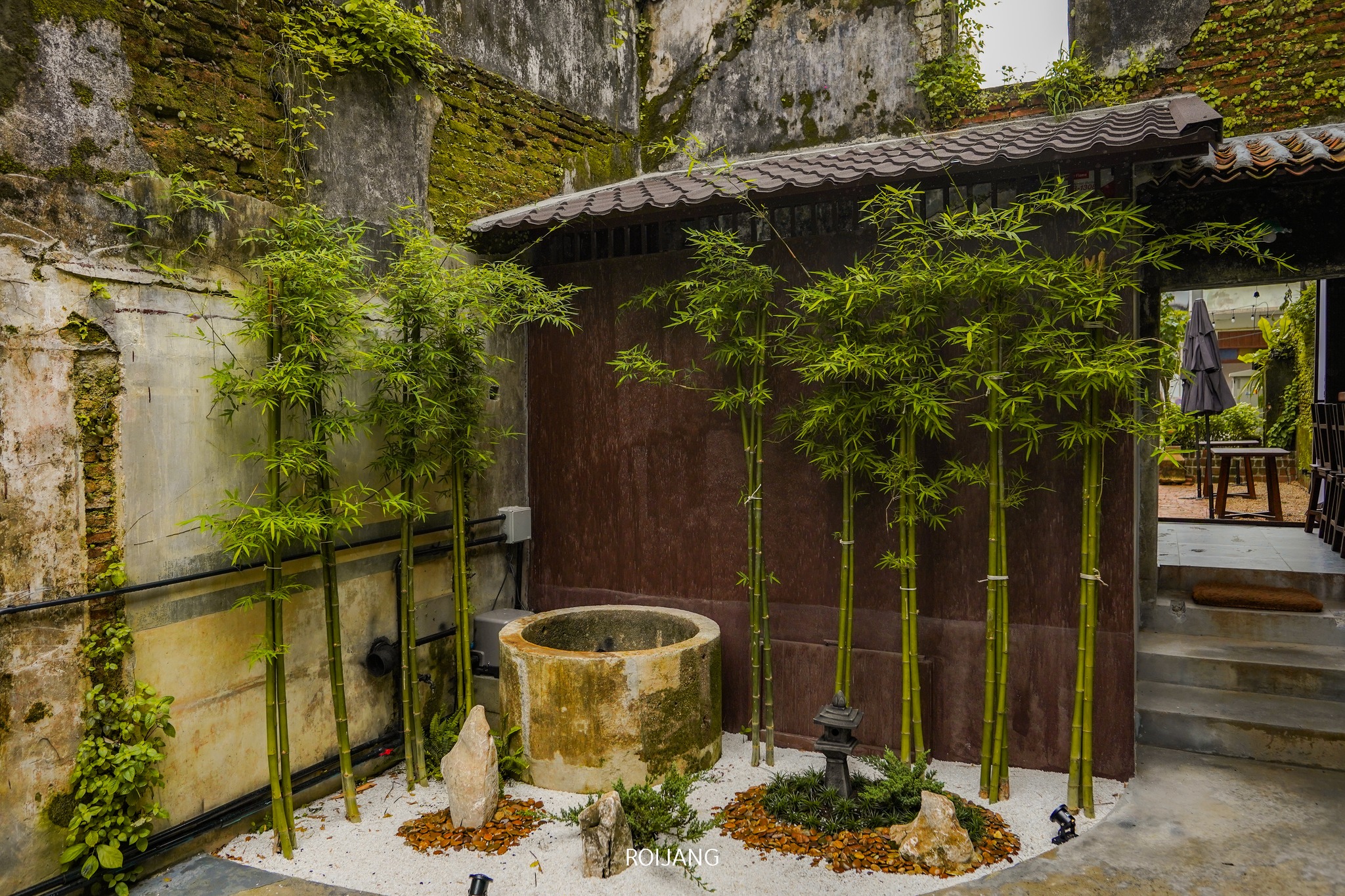 สวนไผ่เล็ก ๆ และบ่อน้ำหินในลานบ้านที่มีผนังเก่าแก่ กำแพงกรวด และทางเดินมีหลังคาในพื้นหลัง—ภาพชวนมองของเสน่ห์เงียบสงบของ ที่เที่ยวตะกั่วป่า