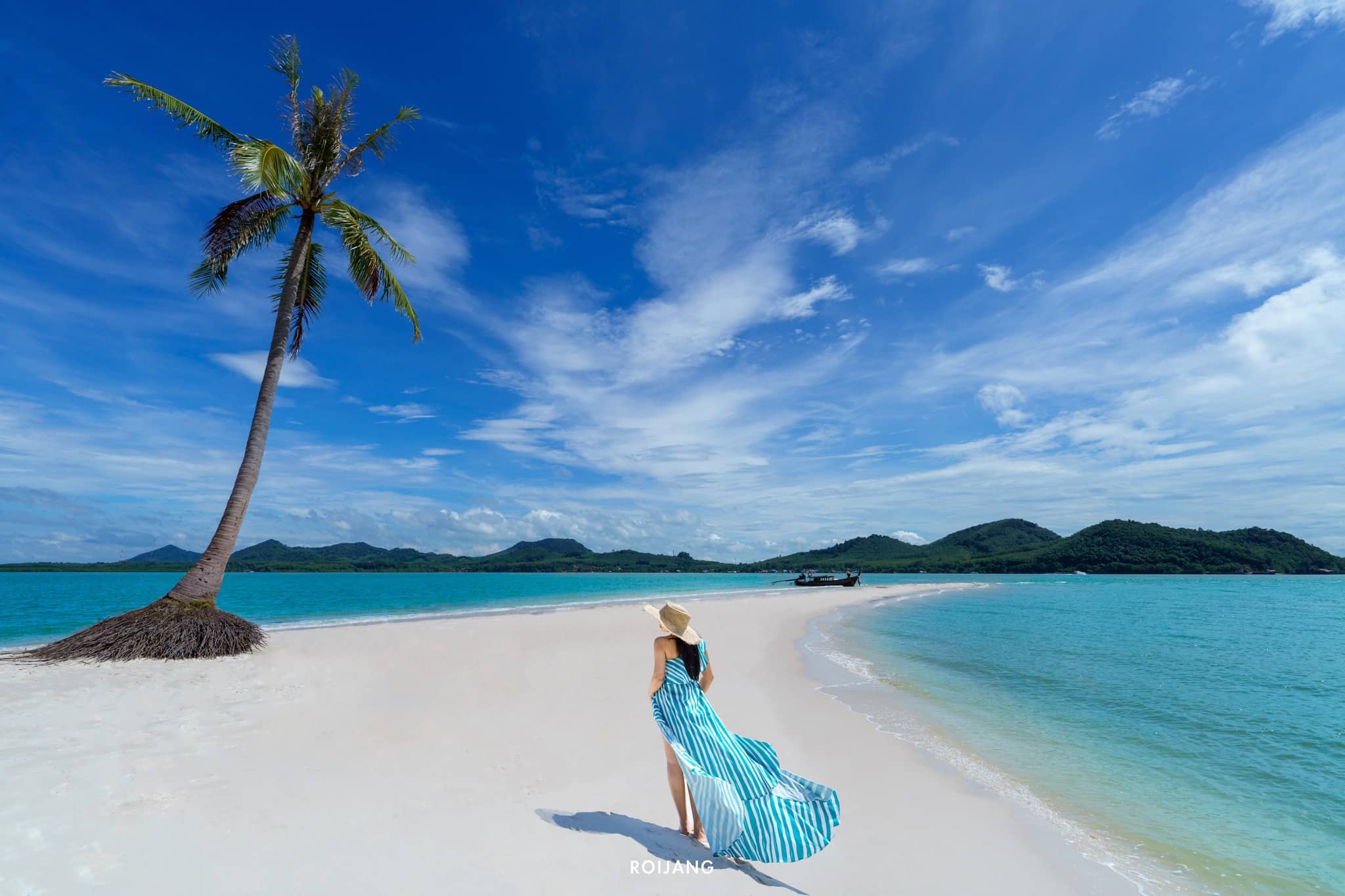 ผู้หญิงในชุดเดรสสีน้ำเงินและหมวกกันแดดยืนอยู่บนชายหาดทรายขาวใกล้ต้นปาล์มที่โค้งงอ มองเห็นน้ำสีฟ้าครามและเนินเขาสีเขียวไกลโพ้นภายใต้ท้องฟ้าสีคราม ณ สถานที่ท่องเที่ยวตะกั่วป่า .