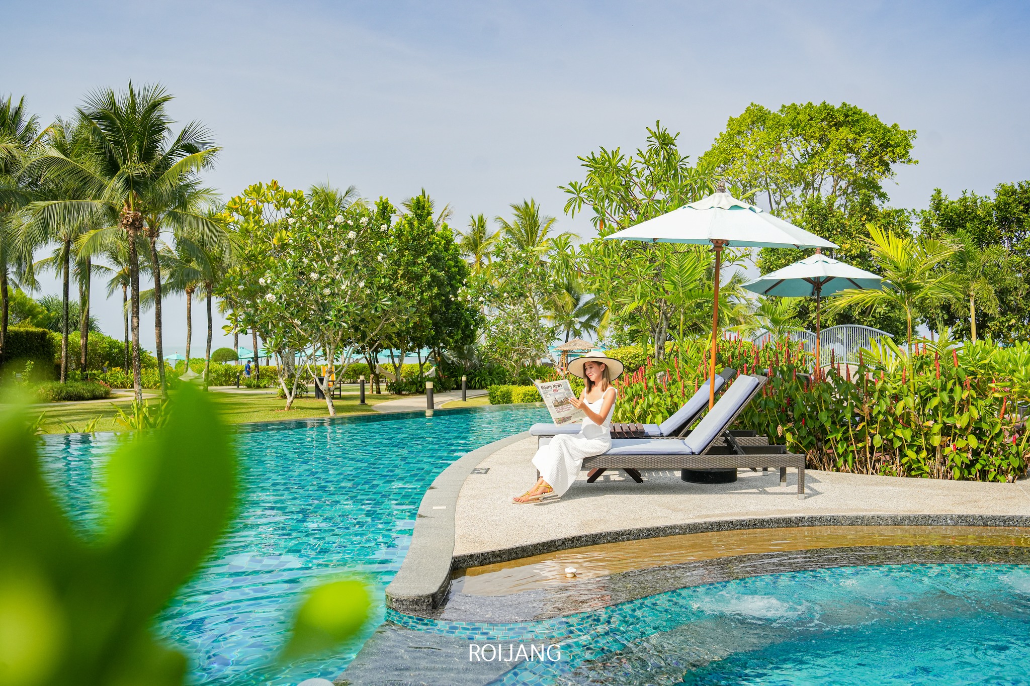 ผู้หญิงในชุดเดรสสีขาวและหมวกกันแดดนั่งอยู่บนเก้าอี้ชายหาดข้างสระว่ายน้ำ กำลังอ่านหนังสือพิมพ์อยู่ ท่ามกลางต้นไม้และร่มเงา—ถ่ายทอดบรรยากาศผ่อนคลายของ ที่เที่ยวตะกั่วป่า ในวันที่มีแดดได้อย่างสมบูรณ์แบบ.