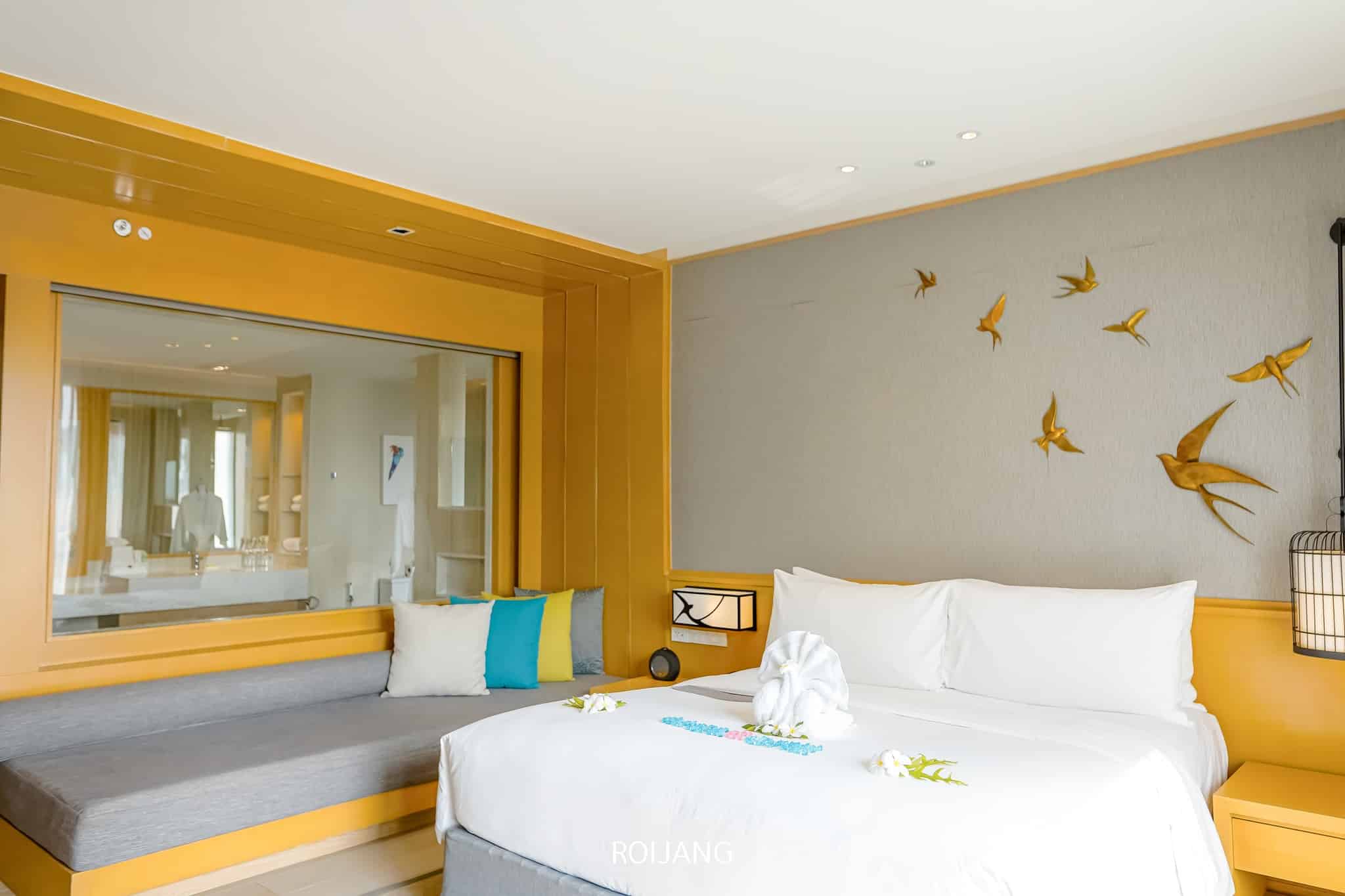 ห้องพักโรงแรมสมัยใหม่ใกล้แหล่ง ท่อง เที่ยวตะกั่วป่า ประกอบด้วยเตียงคู่พร้อมผ้าปู ที่นอนสีขาวสะอาดตา, งานศิลปะพับผ้าเช็ดตัว, ตกแต่งผนังด้วยรูปนก, ม้านั่งมีเบาะ, และหน้าต่างบานใหญ่ที่มองเห็นห้องน้ำ.