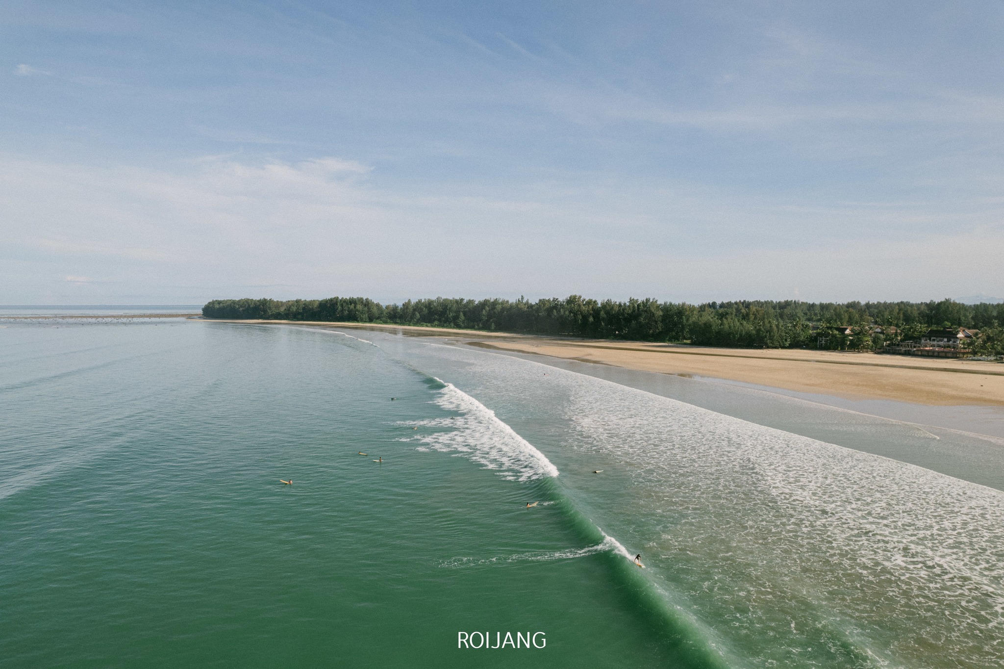 ภาพมุมสูงของชายหาดที่เงียบสงบที่ ที่เที่ยวตะกั่วป่า แสดงให้เห็นคลื่นที่อ่อนโยน นักโต้คลื่นในน้ำ และชายฝั่งที่เรียงรายไปด้วยต้นไม้ภายใต้ท้องฟ้าที่แจ่มใส.