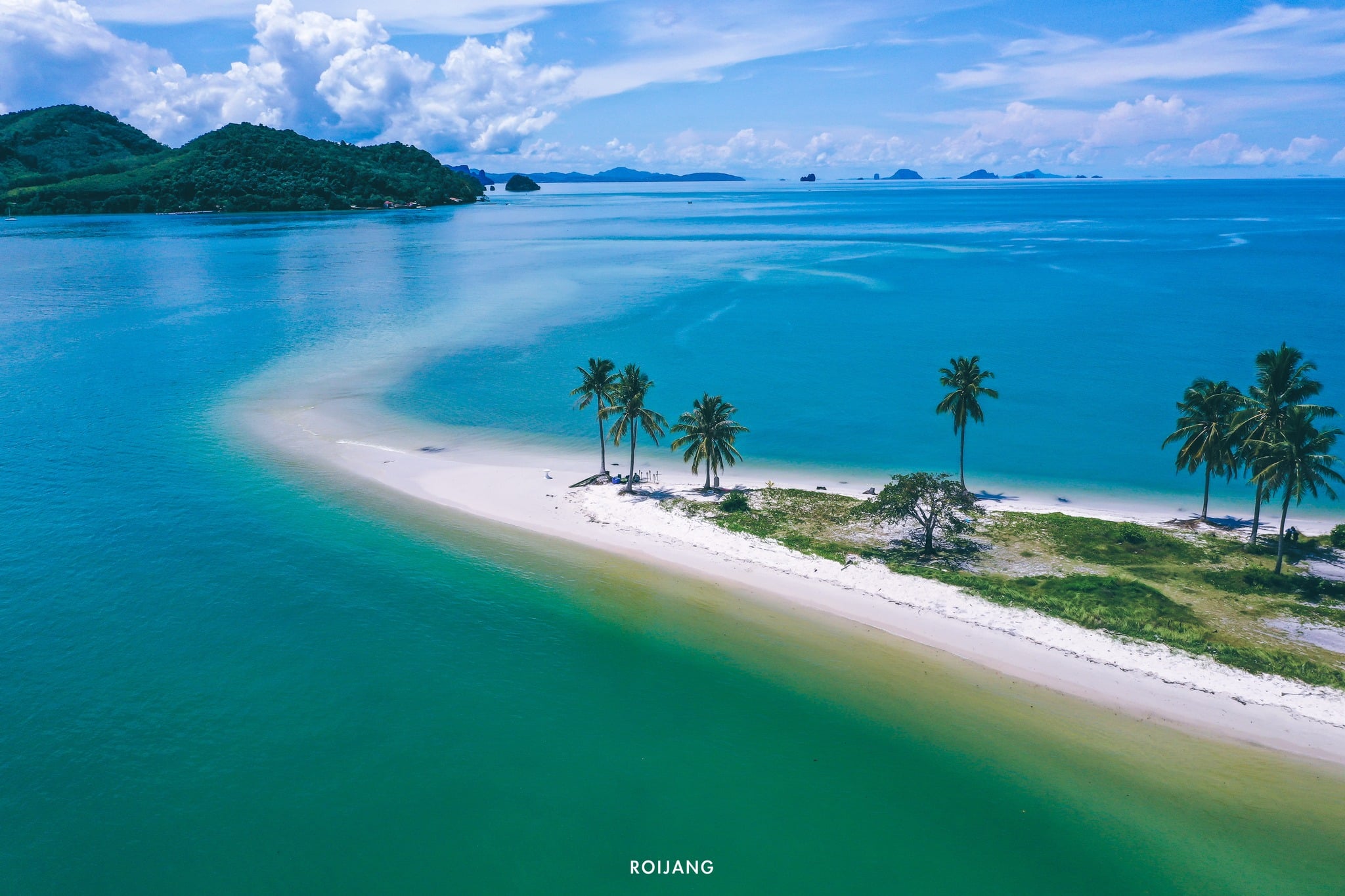แนวชายหาดแคบที่เต็มไปด้วยทรายขาวละเอียดและต้นปาล์มทอดยาวสู่ผืนน้ำสีฟ้าใสสงบภายใต้ท้องฟ้าที่มีเมฆบางส่วน ทำให้ที่นี่เป็นจุดหมายปลายทางที่เงียบสงบใน เที่ยวตะกั่วป่า
