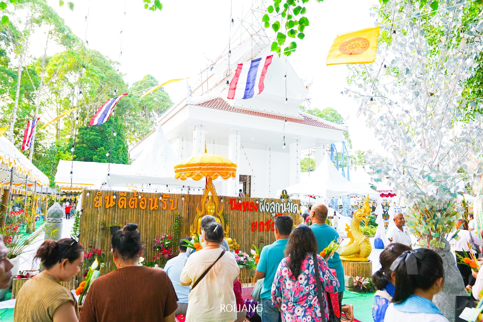กลุ่มคนรวมตัวกันอยู่ด้านนอกวัดไทยที่ประดับประดาด้วยธง รูปปั้น และป้ายต้อนรับ ในระหว่างเทศกาลหรืองานพิธีทางศาสนา—หนึ่งในภาพบรรยากาศที่มีชีวิตชีวาของ ที่เที่ยวตะกั่วป่า