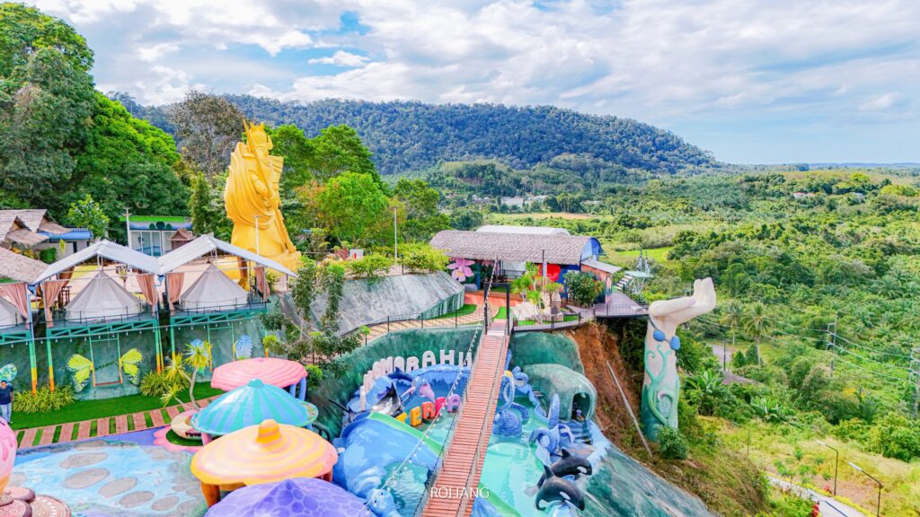 สวนสาธารณะกลางแจ้งสีสันสดใสในสถานที่ท่องเที่ยวตะกั่วป่า พร้อมรูปปั้น เต็นท์ และสะพาน ตั้งอยู่บนเนินเขาที่ล้อมรอบด้วยป่าเขียวชอุ่มและภูเขาภายใต้ท้องฟ้าที่มีเมฆบางส่วน.