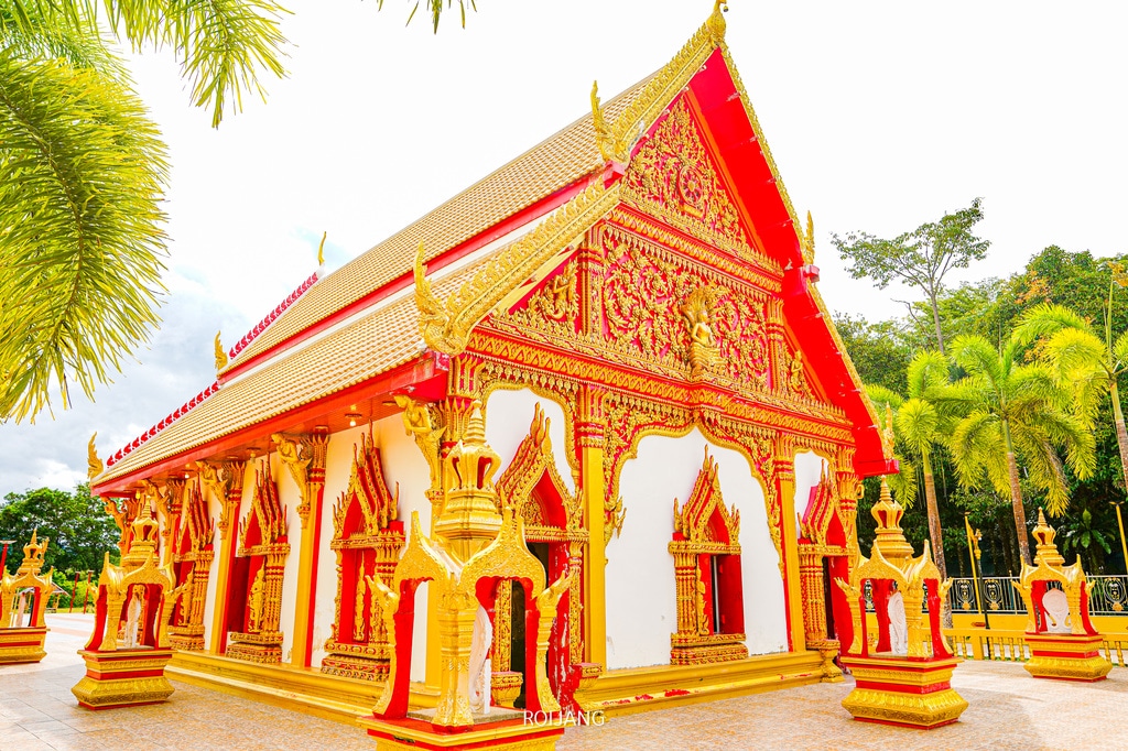 วัดไทยที่เต็มไปด้วยชีวิตชีวา ประดับประดาด้วยลวดลายสีแดงและทองอันวิจิตร ตั้งอยู่ท่ามกลางต้นปาล์มและพืชพรรณเขียวขจีภายใต้ท้องฟ้าสดใส—หนึ่งใน สถานที่ท่องเที่ยวตะกั่วป่า ที่ต้องไปเยือนในตะกั่วป่า.