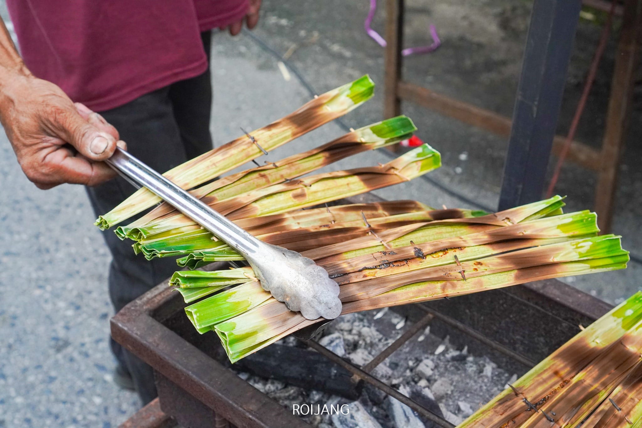 คนกำลังย่างอาหารที่ห่อด้วยใบไม้สีเขียวบนถ่านไฟ โดยใช้คีมโลหะพลิกห่ออาหาร—ภาพที่คุณอาจพบได้ขณะสำรวจสถานที่ท่อง เที่ยวที่ตะกั่วป่า