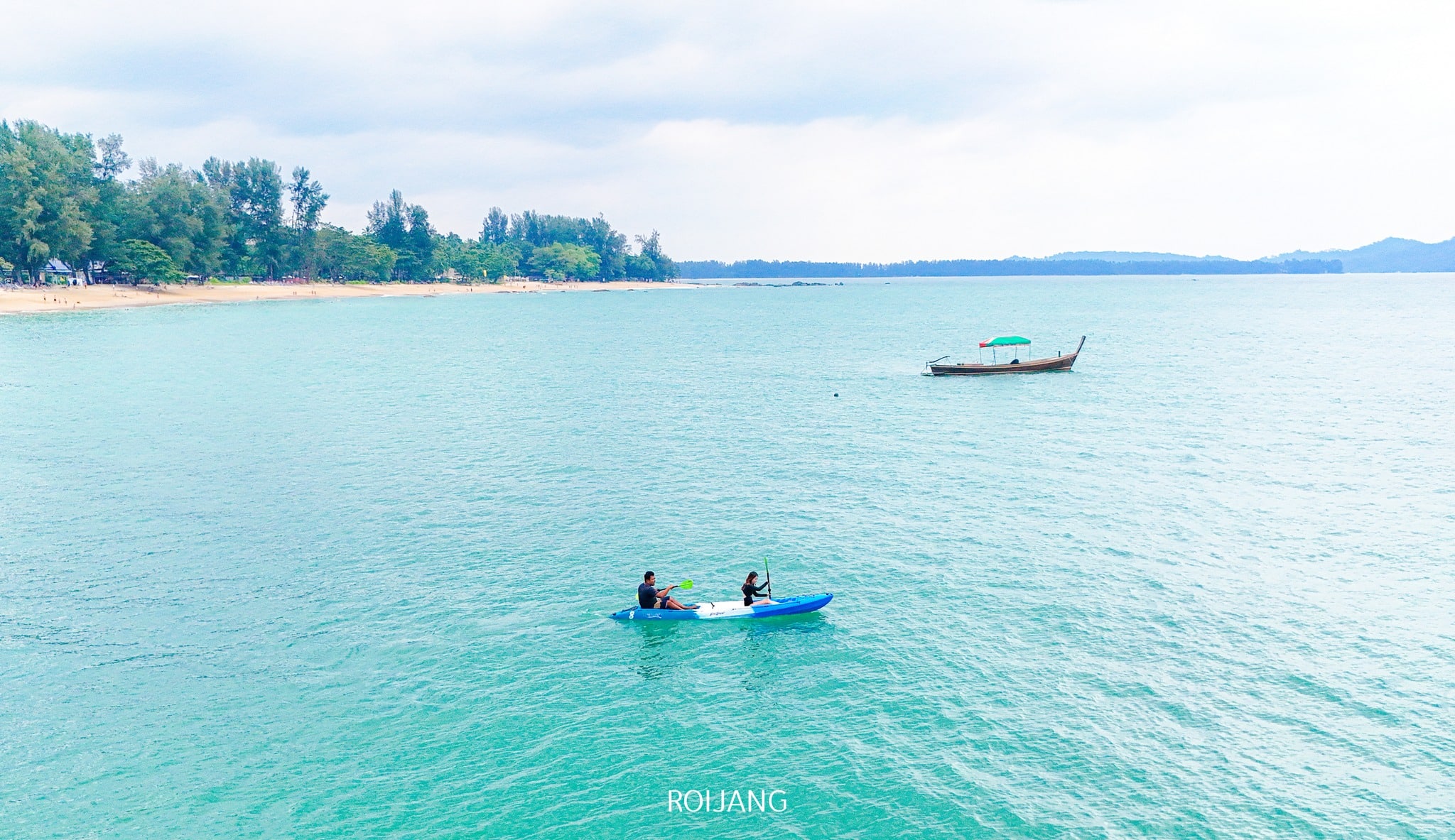 สองคนกำลังพายเรือคายัคสีน้ำเงินบนผืนน้ำสีฟ้าเทอร์ควอยซ์ที่สงบใกล้ชายหาดที่ ที่เที่ยวตะกั่วป่า โดยมีเรือลำเล็กจอดทอดสมออยู่ใกล้ๆ และต้นไม้เรียงรายตามแนวชายฝั่งอันงดงาม.
