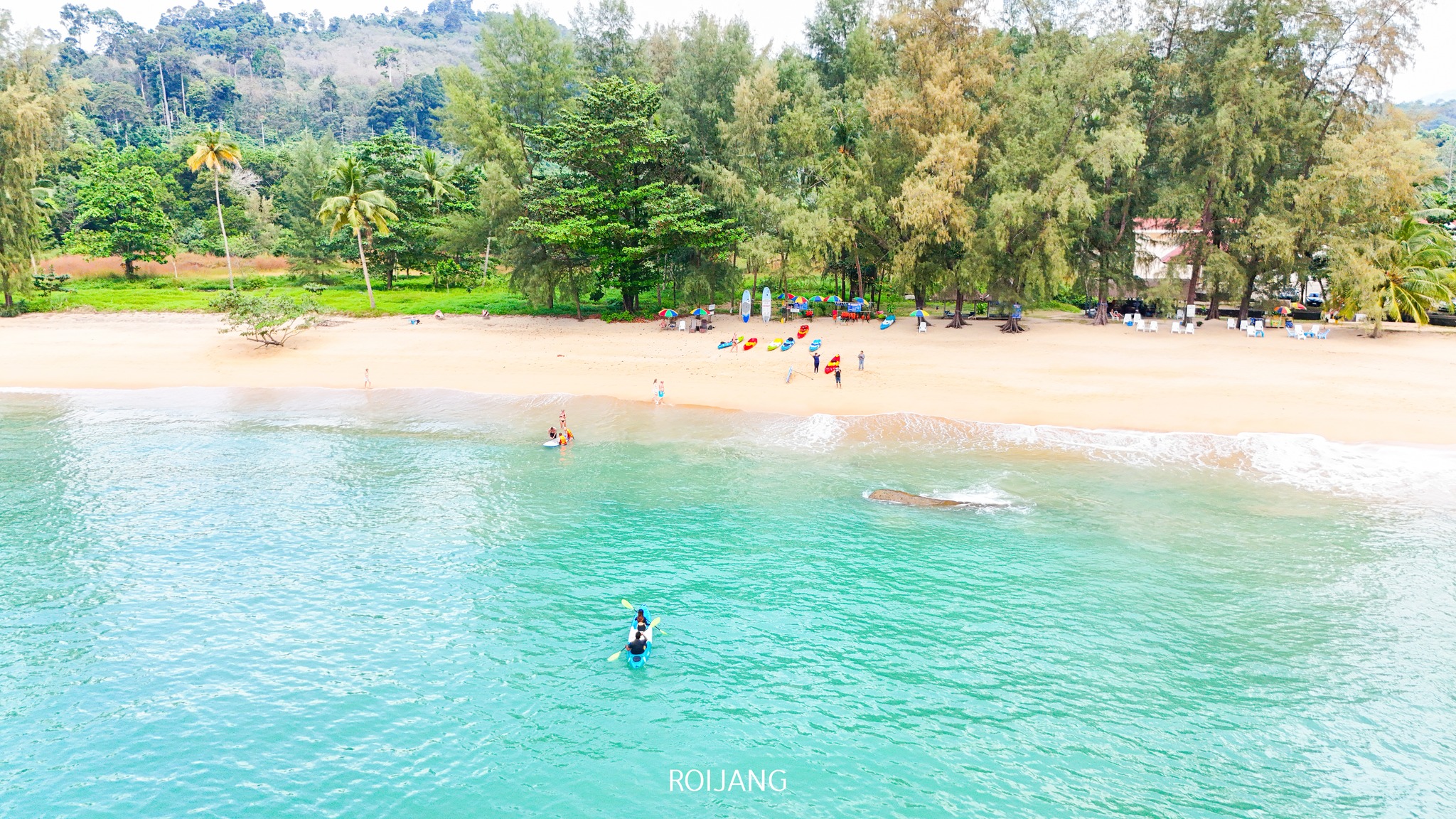 ภาพมุมสูงของชายหาดทรายที่ ท่องเที่ยวตะกั่วป่า มีผู้คนอยู่ใกล้ชายฝั่ง มีเรือคายัคบนทราย และน้ำสีเขียวอมฟ้าที่สงบอยู่เบื้องหน้า.