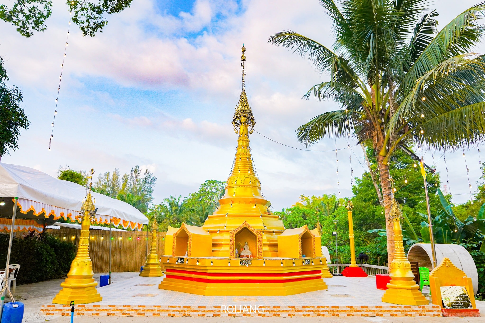 เจดีย์ทองคำประดับลวดลายวิจิตรตั้งตระหง่านอยู่ในพื้นที่กลางแจ้ง รายล้อมด้วยต้นปาล์มและพืชพรรณเขียวขจี ภายใต้ท้องฟ้าที่มีเมฆบางส่วนปกคลุม ทำให้ที่นี่เป็นสถานที่ท่องเที่ยวที่ต้องไปเยือนใน ที่เที่ยวตะกั่วป่า สำหรับผู้ที่แสวงหาความงดงามทางวัฒนธรรม.