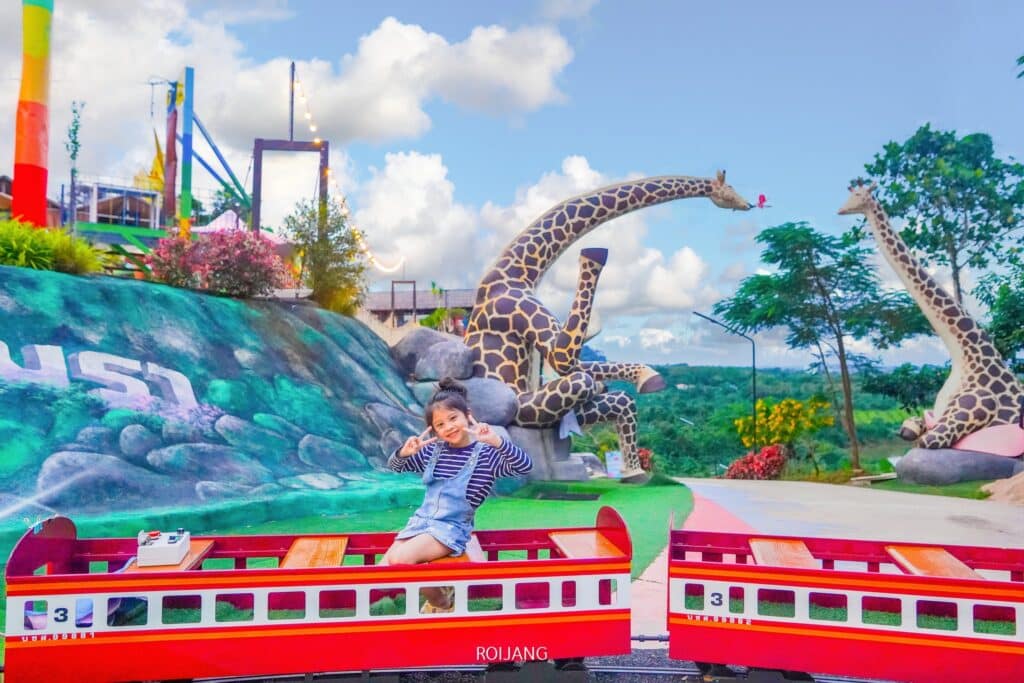 เด็กคนหนึ่งนั่งและโพสท่าบนรถไฟขนาดเล็กสีแดงและขาวที่สวนสาธารณะกลางแจ้งในสถานที่ท่องเที่ยวตะกั่วป่า โดยมีรูปปั้นยีราฟขนาดใหญ่และทิวทัศน์สีสันสดใสเป็นฉากหลังที่สนุกสนาน.