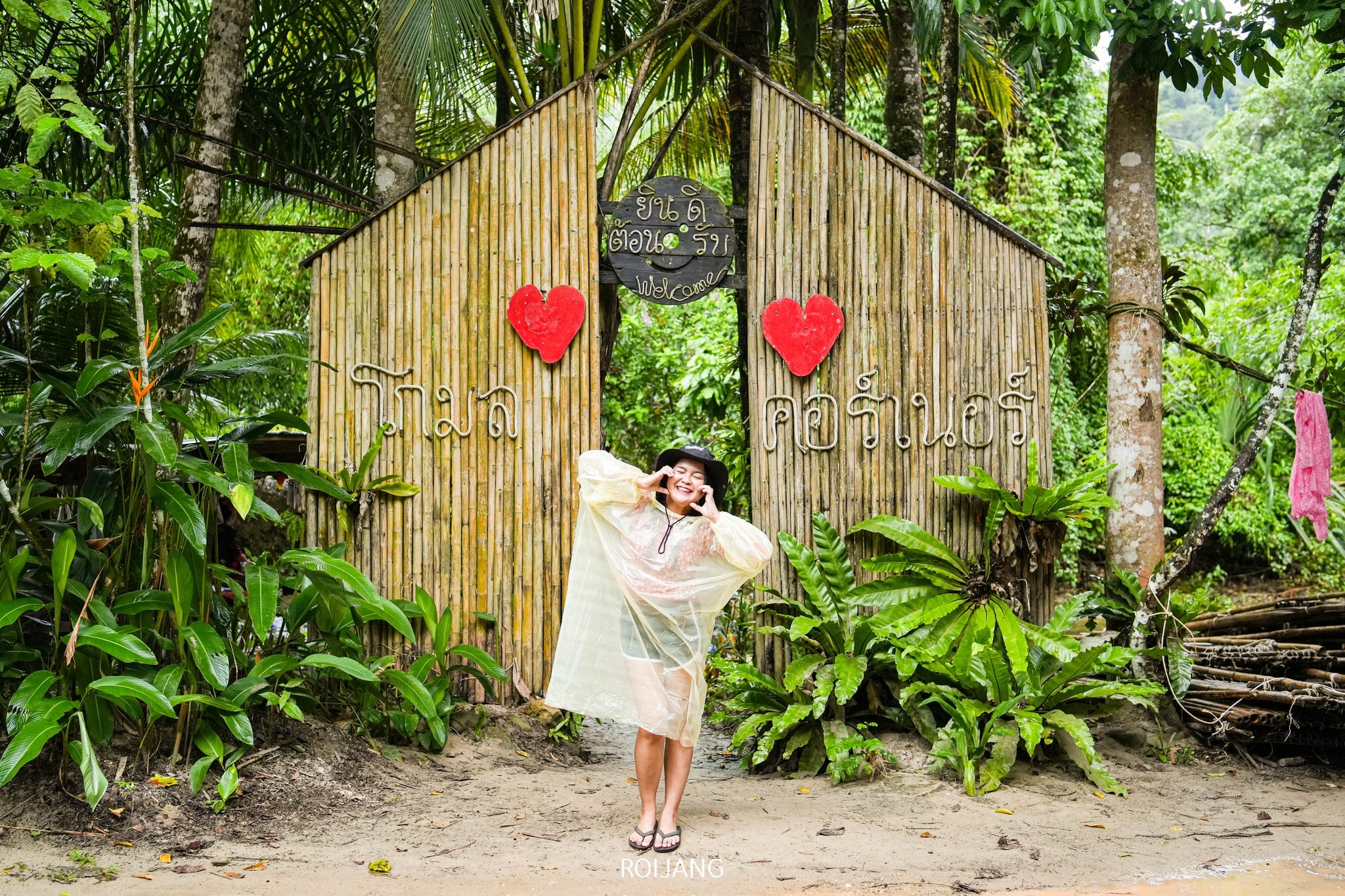บุคคลหนึ่งสวมเสื้อกันฝนและหมวกใสยืนอยู่หน้าประตูไม้ไผ่ที่มีรูปหัวใจสีแดงสองดวงและตัวอักษรภาษาไทย ล้อมรอบด้วยพืชสีเขียวชอุ่ม—เป็นภาพที่น่าดึงดูดใจที่ ที่เที่ยวตะกั่วป่า.