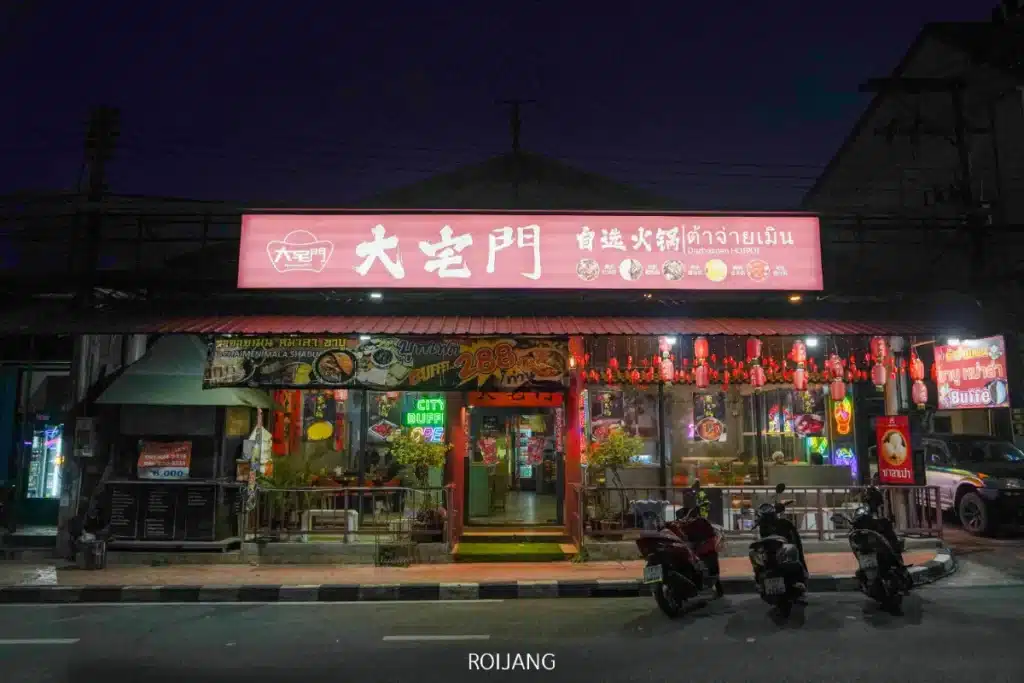 ร้านอาหารเอเชียที่สว่างไสวในยามค่ำคืน มีโคมไฟสีแดง ป้ายไฟนีออน และรถจักรยานยนต์จอดอยู่ด้านหน้า พร้อมชื่อ "ต้าจ่ายเมิน" ส่องแสงสว่างอยู่เหนือทางเข้า.