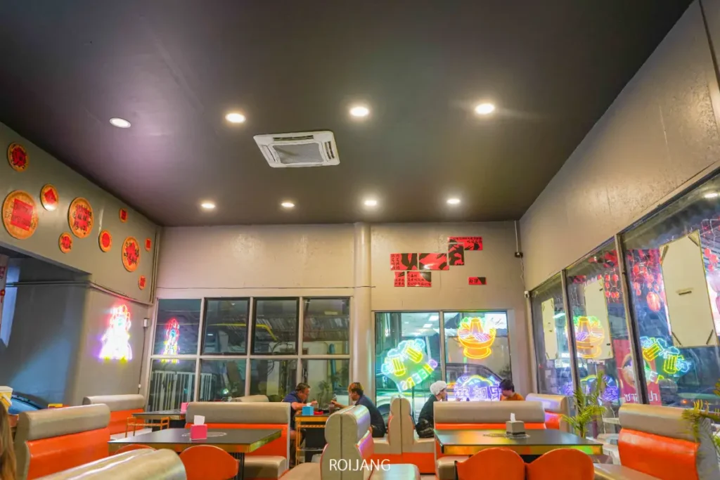 ภายในร้านอาหารที่สว่างไสว ตกแต่งด้วยสไตล์โมเดิร์น มีป้ายไฟนีออนติดอยู่บนผนังและหน้าต่าง ผู้คนนั่งอยู่ที่โต๊ะบูธ และมีป้ายไฟ "ต้าจ่ายเมิน" ส่องแสงสว่างอยู่เหนือทางเข้า.