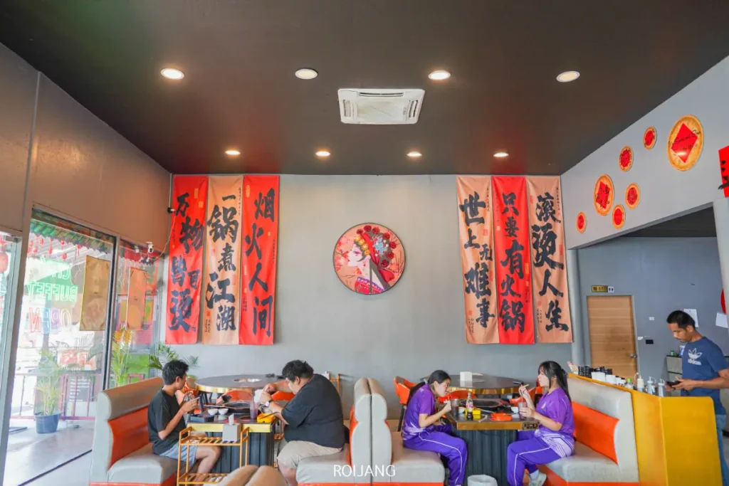 ผู้คนรับประทานอาหารที่ร้านอาหารเอเชียสมัยใหม่ซึ่งมีป้ายผนังสีแดงและดำที่มีตัวอักษรจีน, การตกแต่งผนังเป็นวงกลม, และลวดลาย ต้าจ่ายเมิน ที่โดดเด่นช่วยเสริมบรรยากาศ.
