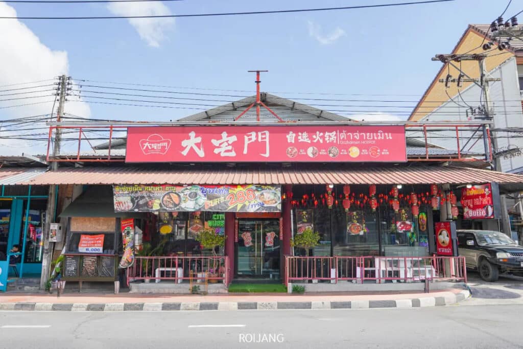 ภาพถนนหน้าร้านอาหาร ต้าจ่ายเมิน มีป้ายสีแดงเขียนเป็นภาษาไทยและภาษาจีน ตกแต่งด้วยโคมไฟสีแดงและแบนเนอร์ ตั้งอยู่ติดถนนที่มีรถยนต์จอดอยู่.