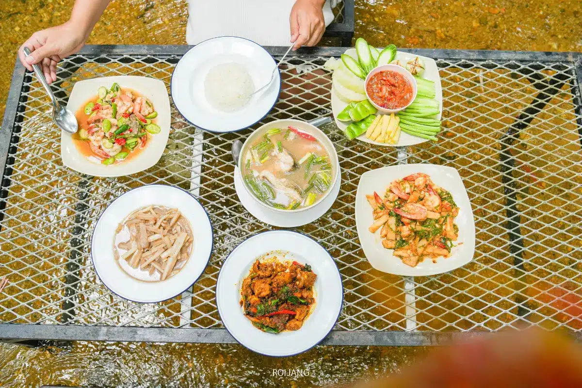 โต๊ะที่ร้านมีสุข ท้ายเหมือง ตั้งอยู่เหนือผืนน้ำ มีอาหารไทยจัดแสดงอยู่ เช่น ข้าว ซุป ผัก กุ้ง และเนื้อ พร้อมด้วยมือที่ถือช้อนเหนือจาน.