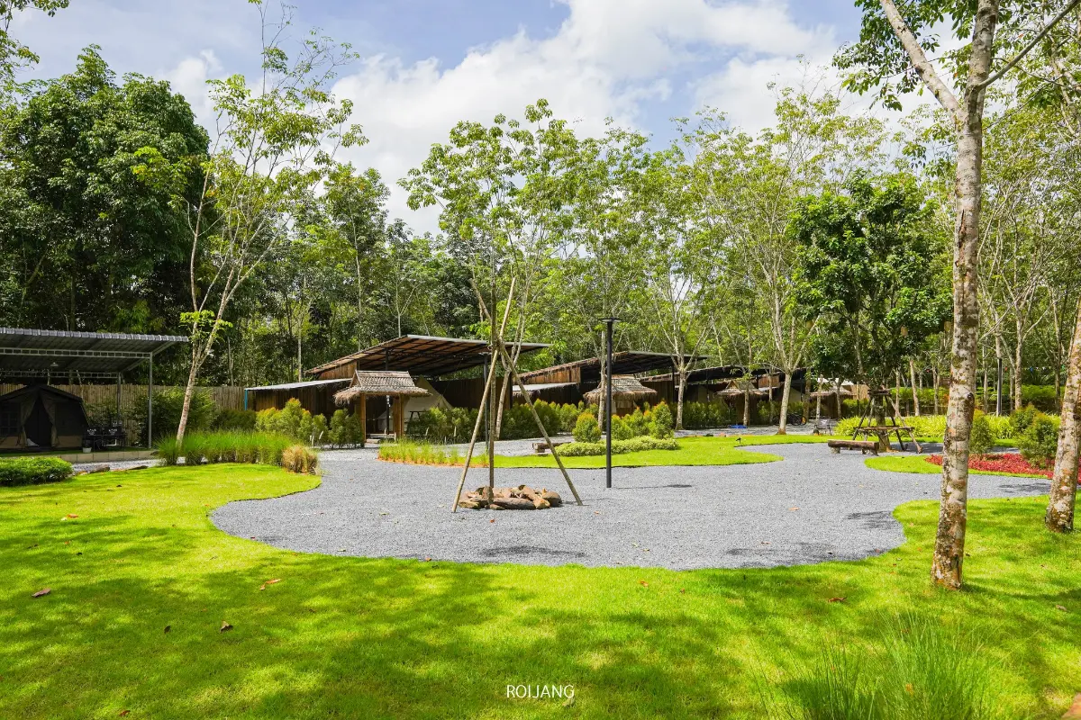 พื้นที่กลางแจ้งที่จัดภูมิทัศน์อย่างสวยงามที่ มีสุข ท้ายเหมือง ประกอบด้วยสนามหญ้าสีเขียว ทางเดินกรวด ต้นไม้ และโครงสร้างไม้ ภายใต้ท้องฟ้าที่มีเมฆบางส่วน.