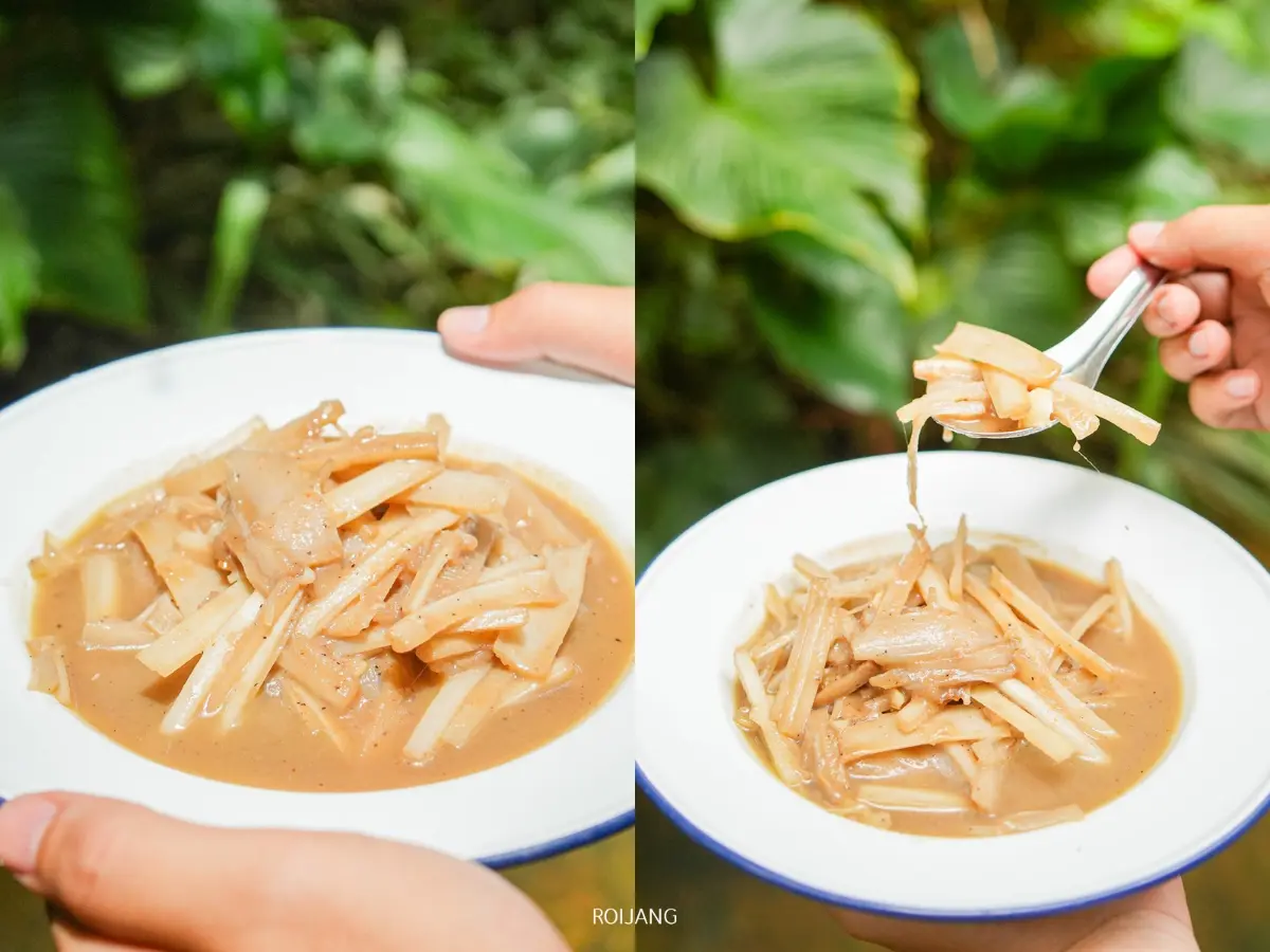 บุคคลหนึ่งถือจานที่มีหน่อไม้ในซอสสีน้ำตาลที่ร้านมีสุข ท้ายเหมือง โดยมีภาพหนึ่งที่แสดงส้อมกำลังยกชิ้นหน่อไม้ขึ้น มีพืชใบเขียวอยู่ในพื้นหลังที่เบลอ.