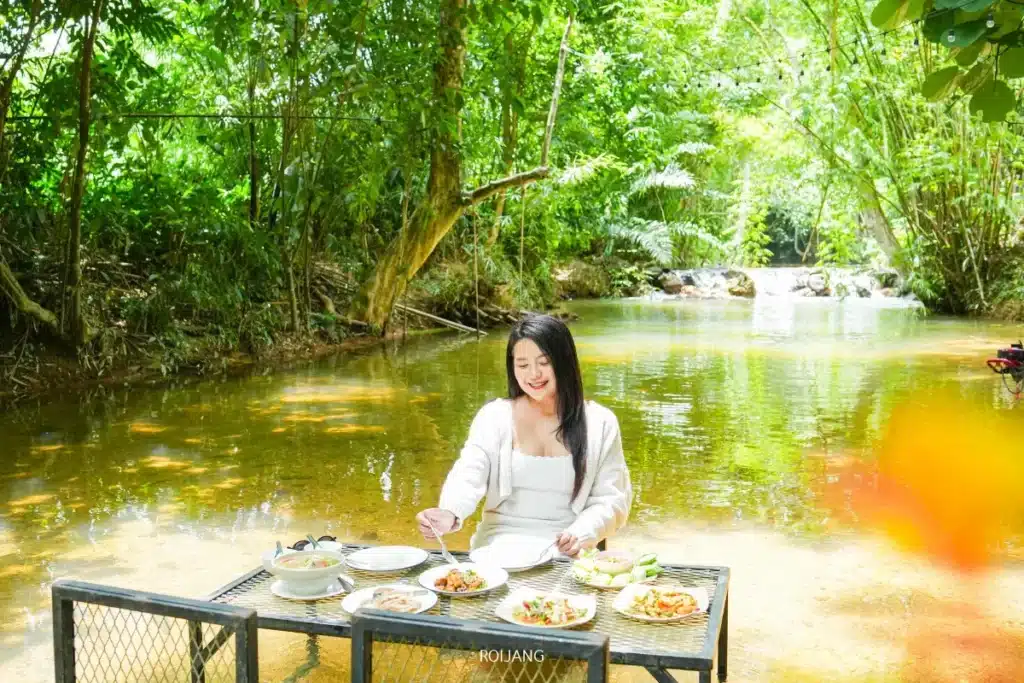 ผู้หญิงคนหนึ่งนั่งอยู่ที่โต๊ะซึ่งจัดวางด้วยอาหารหลากหลายชนิดในลำธารตื้นใสที่ ศิ-วรา คาเฟ่ ล้อมรอบด้วยต้นไม้และพืชพรรณเขียวชอุ่ม.