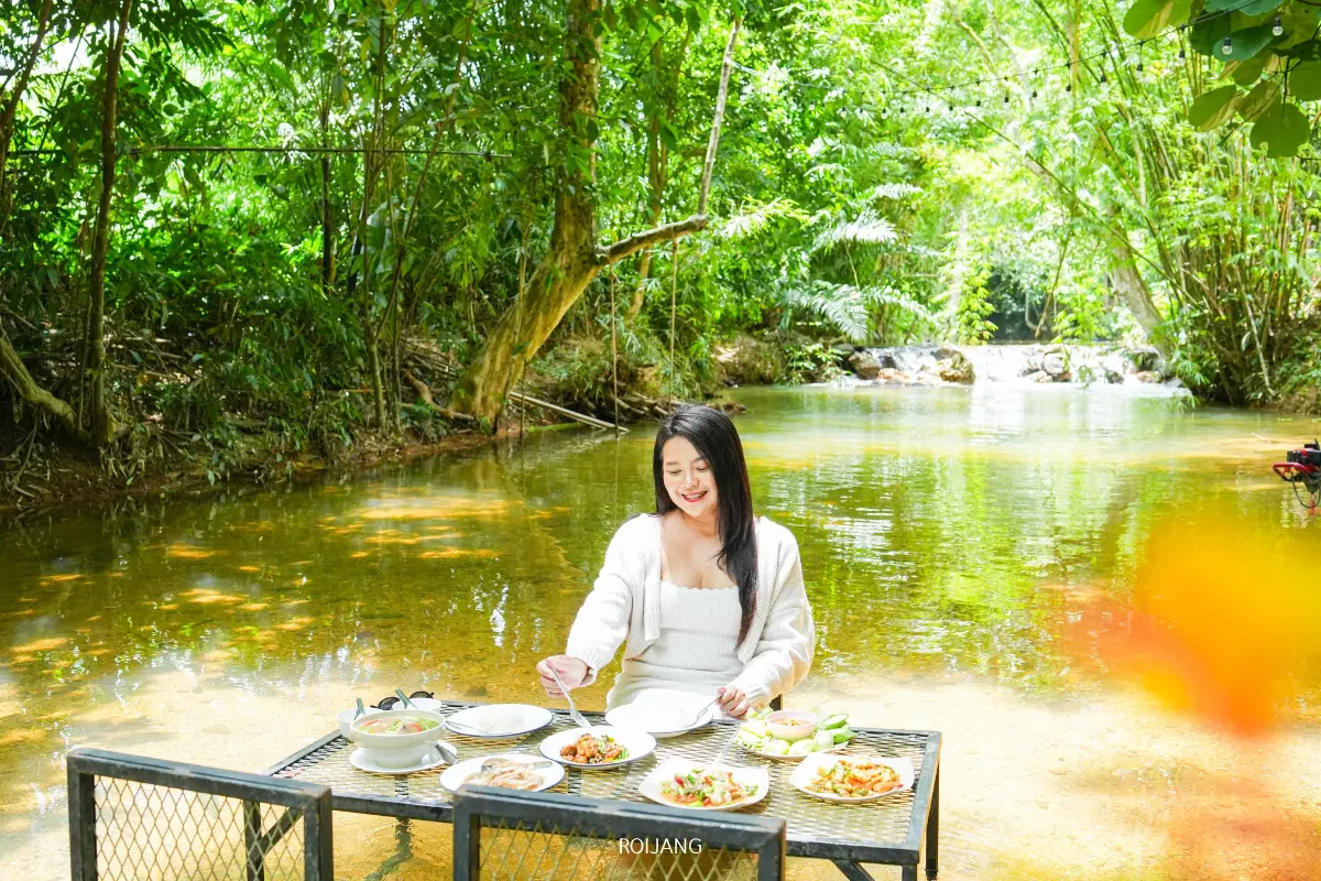ผู้หญิงคนหนึ่งนั่งอยู่ที่โต๊ะซึ่งจัดวางด้วยอาหารหลากหลายชนิดในลำธารตื้นใสที่ มีสุข ท้ายเหมือง ล้อมรอบด้วยต้นไม้และพืชพรรณเขียวชอุ่ม.