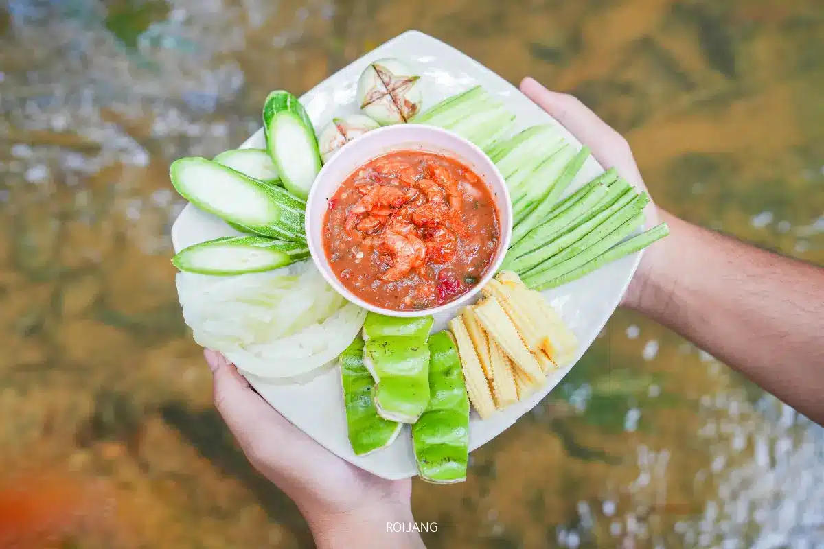 จานผักสดหลากชนิดและข้าวโพดอ่อนจัดวางรอบชามน้ำจิ้มพริกกุ้งที่ยกด้วยสองมือเหนือลำธารที่ มีสุข ท้ายเหมือง.