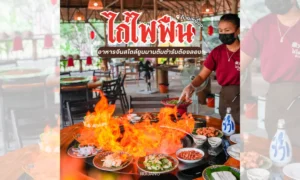 พนักงานร้านอาหารสวมหน้ากากกำลังเสิร์ฟอาหารบนโต๊ะที่มีเปลวไฟลุกโชนจากจานไก่ไฟฟืน มีอาหารไทยหลากหลายชนิดจัดแสดงอยู่ และลูกค้าต่างนั่งรับประทานอาหารอยู่ด้านหลัง.