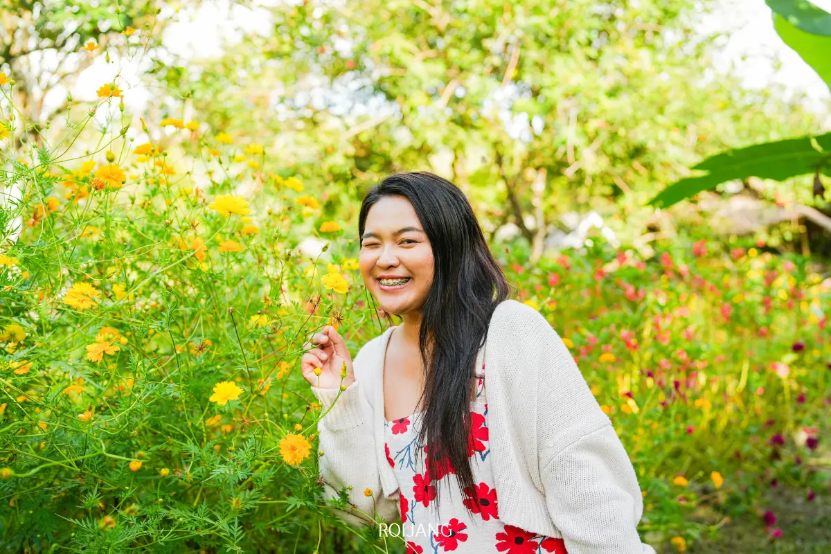 ผู้หญิงคนหนึ่งที่มีผมยาวสีดำ สวมเสื้อสเวตเตอร์สีขาวทับชุดเดรสลายดอกไม้ ยืนยิ้มอยู่ในสวนที่มีดอกไม้สีเหลืองและสีชมพู ขณะที่เครื่องประดับไก่ไฟฟ้าเพิ่มความมีเสน่ห์ให้กับฉากที่สดใส.