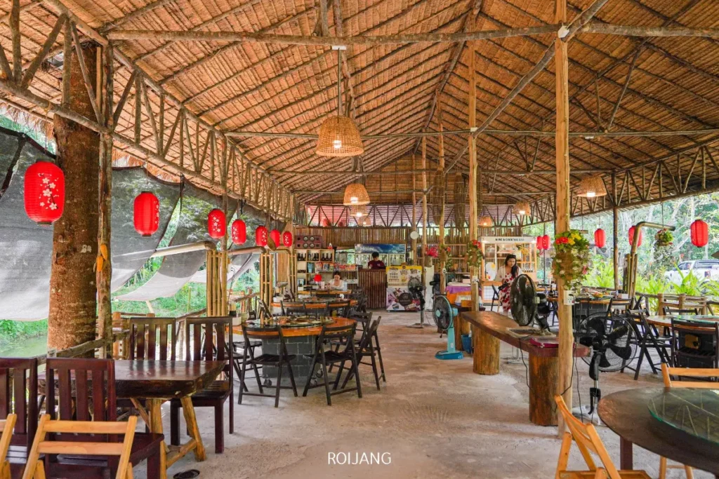 ร้านอาหารกลางแจ้งพร้อมเฟอร์นิเจอร์ไม้ หลังคามุงจากไม้ไผ่ และโคมไฟแขวน ให้แสงธรรมชาติและล้อมรอบด้วยพื้นที่สีเขียว—เหมาะสำหรับการเพลิดเพลินกับอาหารจานเด่นอย่าง ไก่ย่างไฟฟืน ในบรรยากาศที่เงียบสงบ.