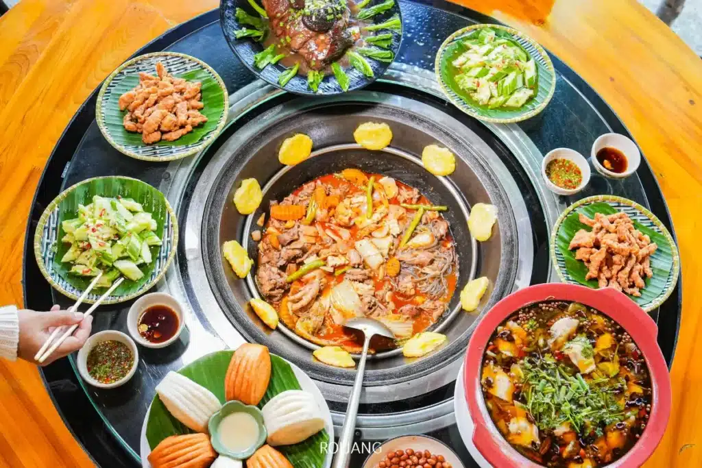 อาหารหลากหลายชนิด รวมถึงหม้อไฟกลางที่มีเนื้อสัตว์ ผัก และไก่ไฟฟ้า ถูกจัดเรียงบนโต๊ะไม้กลมพร้อมจานข้างและน้ำจิ้ม.